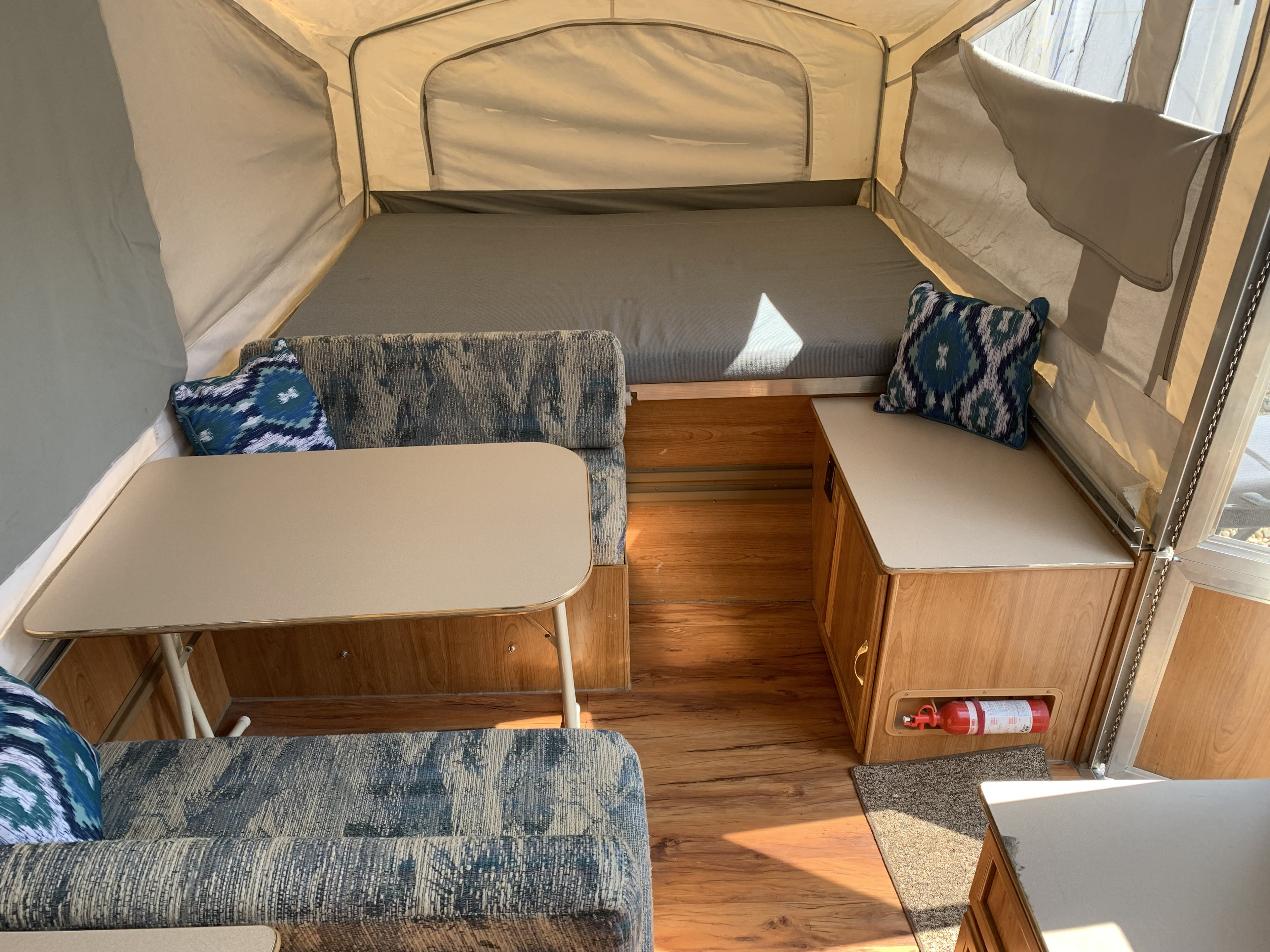 RV rental pop-up camper interior: a cozy bed, dinette area with table, and a safety fire extinguisher are visible on wood flooring.. Forest River Palomino 2003