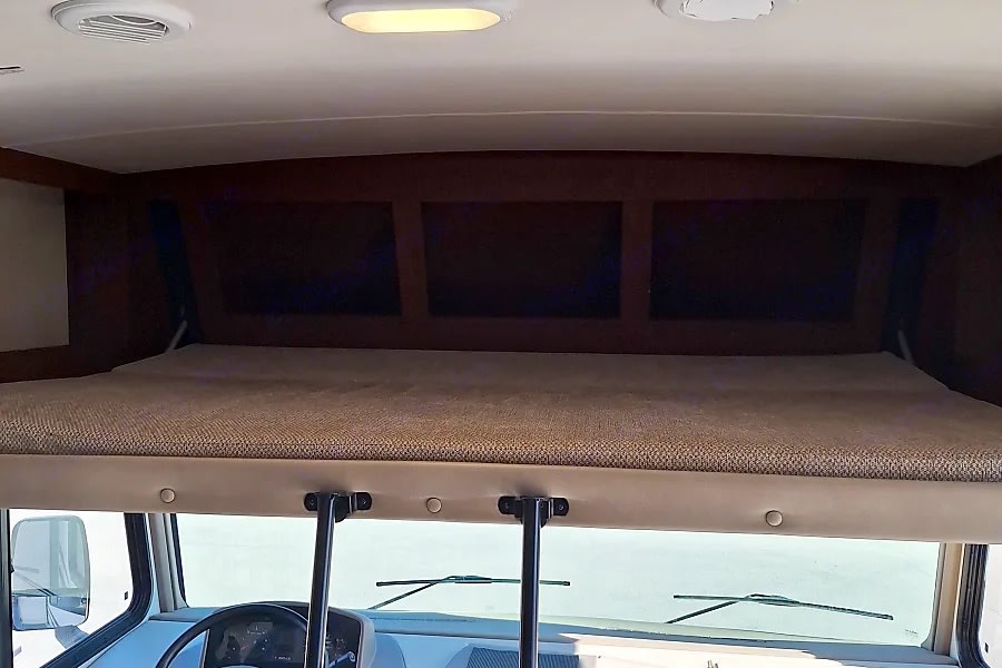 Inside an RV rental, a well-lit cab-over bunk with a brown mattress and three overhead storage compartments.. FR3 FR3 Motorhome 2018