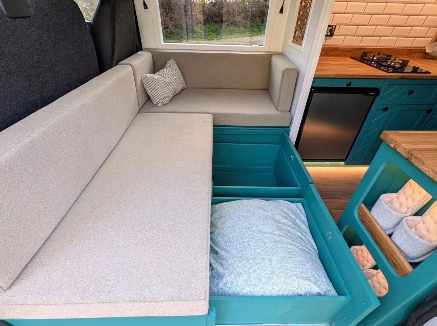 RV rental interior features L-shaped seating that converts to a bed, revealing teal under-seat storage and a compact kitchen.. Peugeot Boxer L4H2 2021