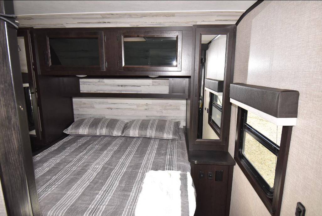 Inside an RV rental: a cozy bedroom with a striped bed, dark wood overhead cabinets, and large windows.. Jayco Jay Feather 2022