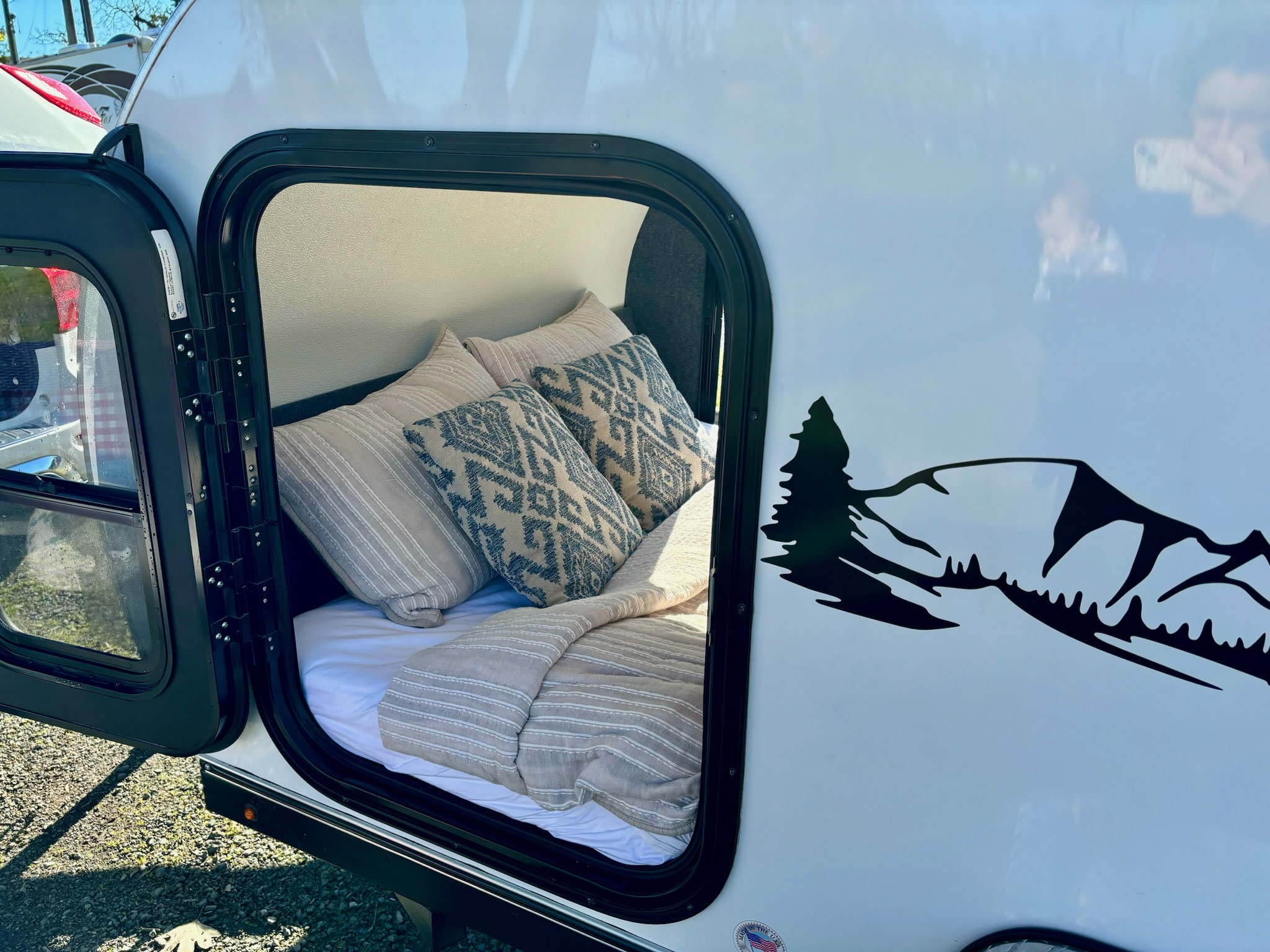 Comfort awaits in this RV rental! Open door shows a cozy bed inside this white travel trailer with a mountain graphic.. American Teardrop Merlin 2021