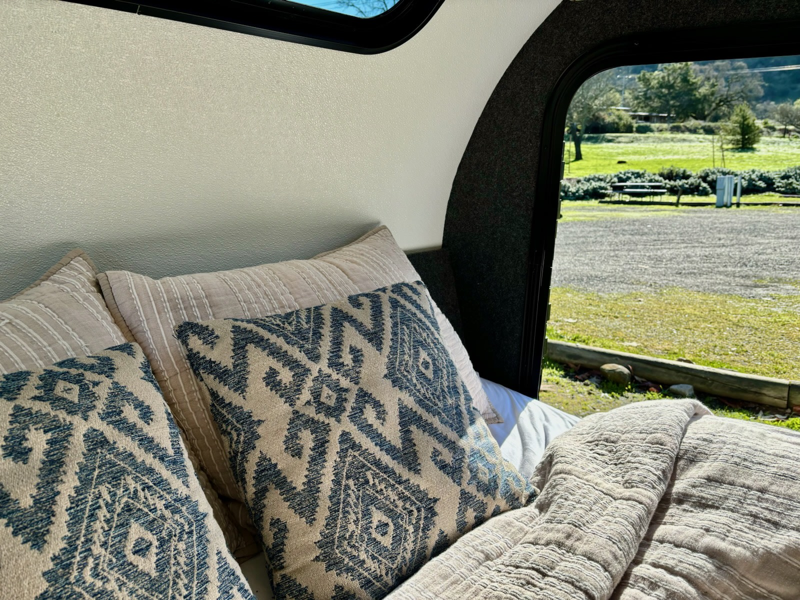 Inviting rv rental bed with textured, patterned pillows, offering a peaceful view of a sunlit outdoor campsite.. American Teardrop Merlin 2021