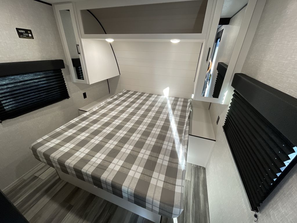 Modern RV rental bedroom: cozy plaid bed, white overhead cabinets, and dark blinds. A compact, comfortable sleeping space.. Jayco 265BHS 2025