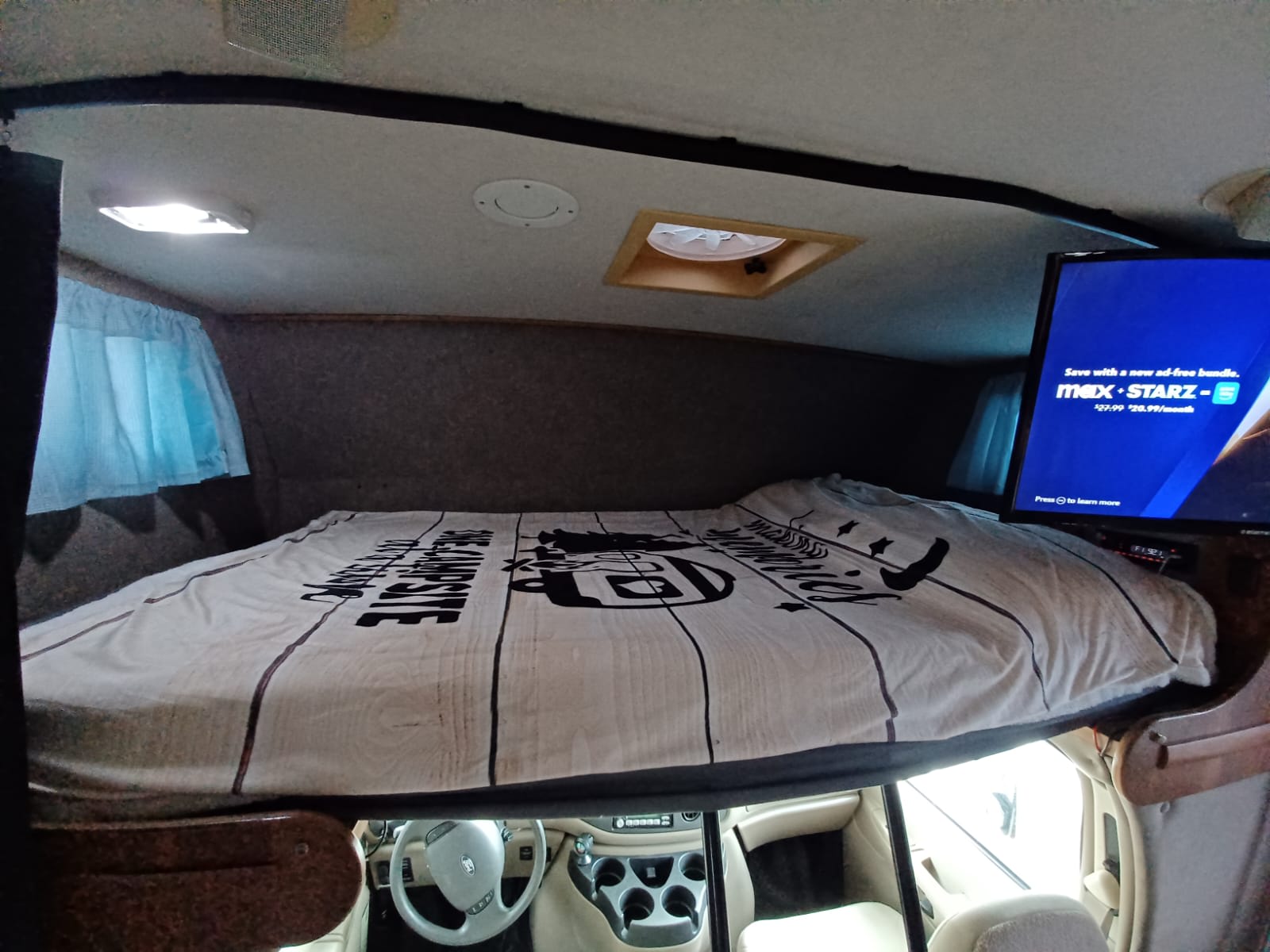RV rental interior: cozy overhead bunk bed above the cab, featuring a TV, ceiling vent, and curtain.. Winnebago Outlook 2007