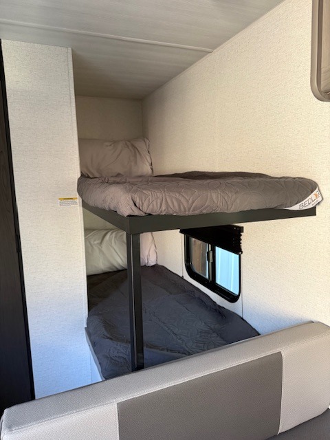 Compact RV rental interior showing stacked bunk beds with grey bedding, a small window, and a light-colored bench.. Keystone Hideout 175BH 2025