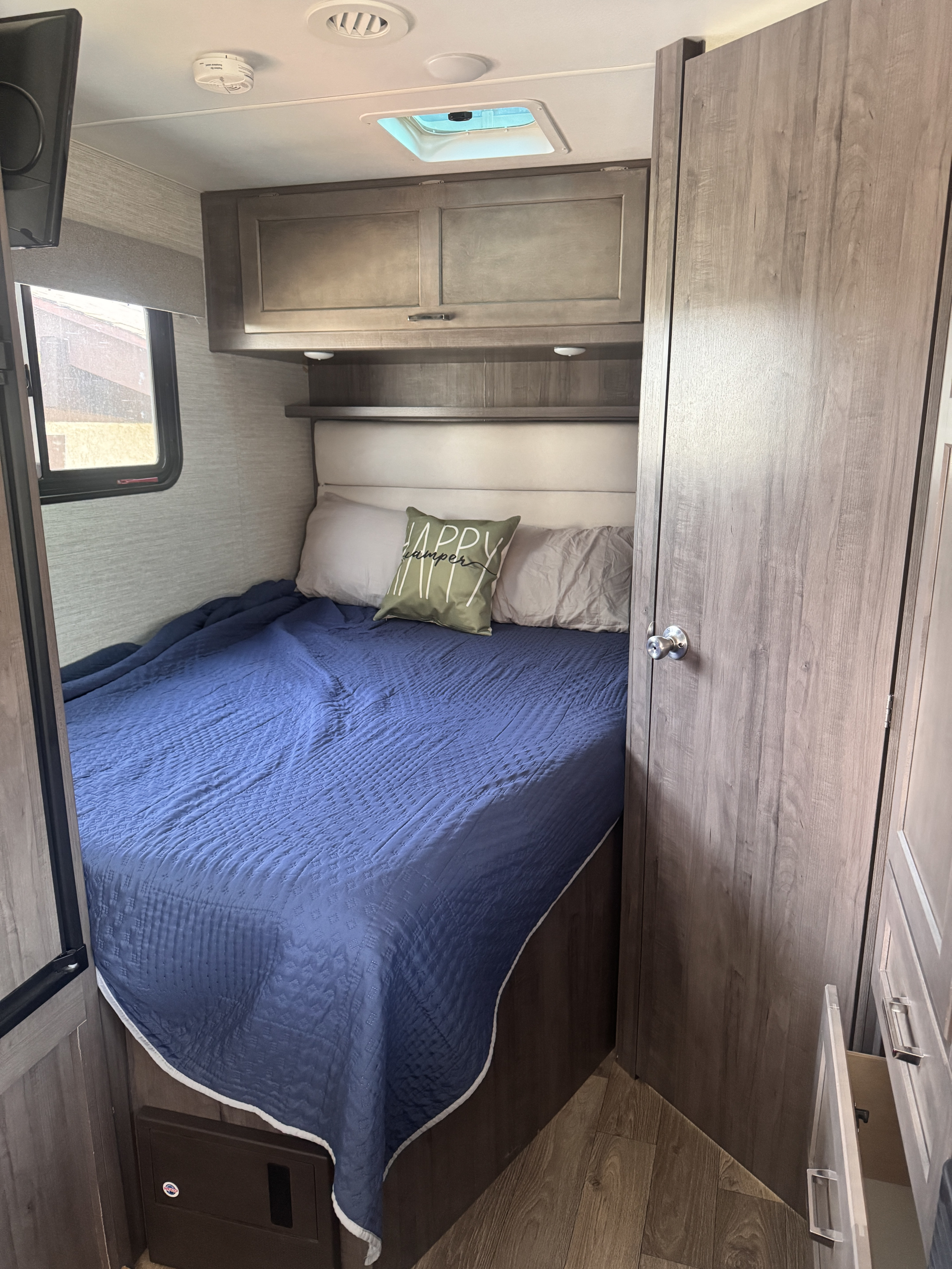 This inviting RV rental bedroom features a comfy bed with a blue comforter, "HAPPY camper" pillow, and built-in storage.. Winnebago Minnie Winnie 2020