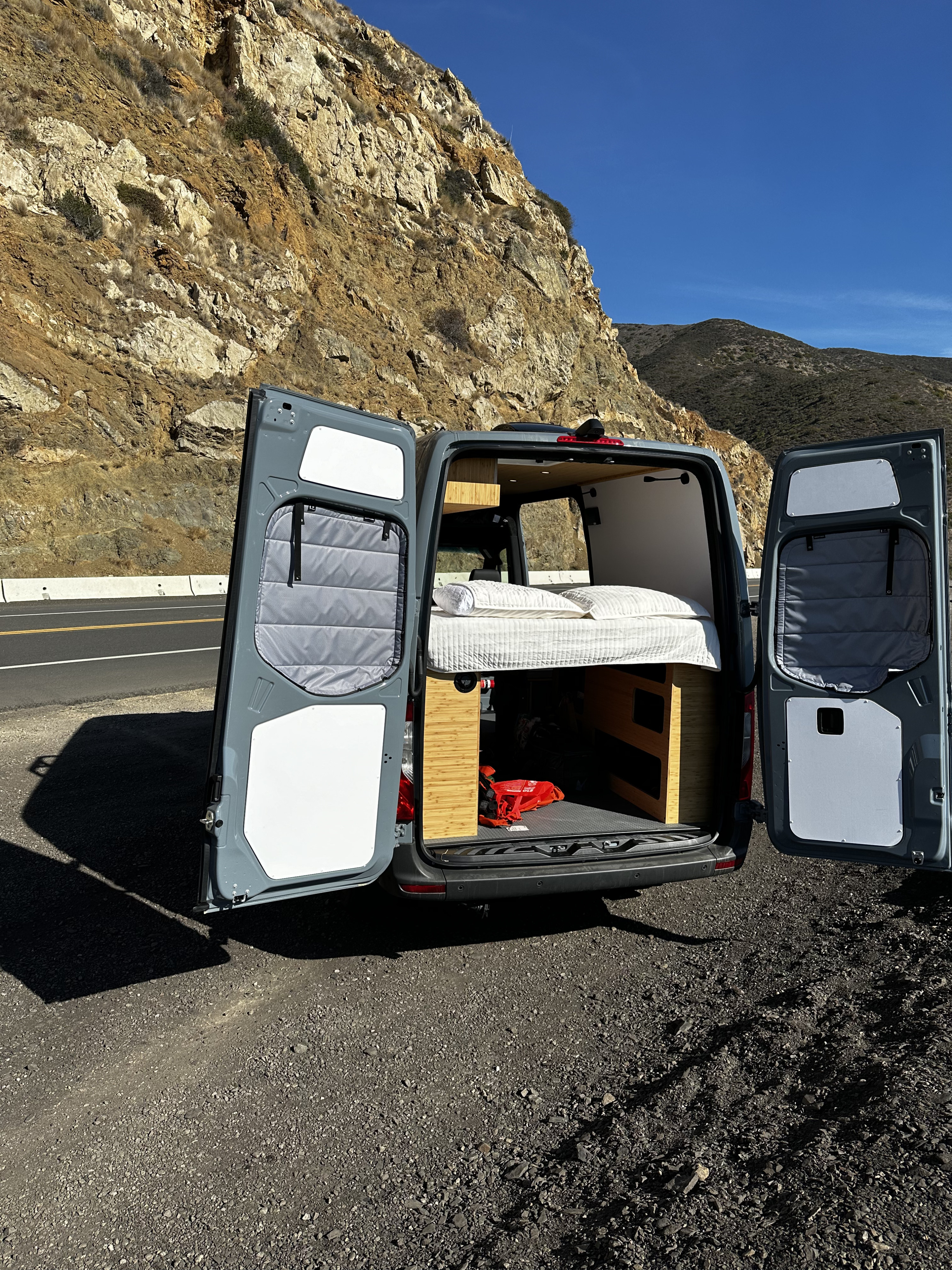 This RV rental van, with its cozy bed revealed, is parked on a scenic mountain road. Perfect for travel adventures!. Mercedes-Benz Sprinter 2024