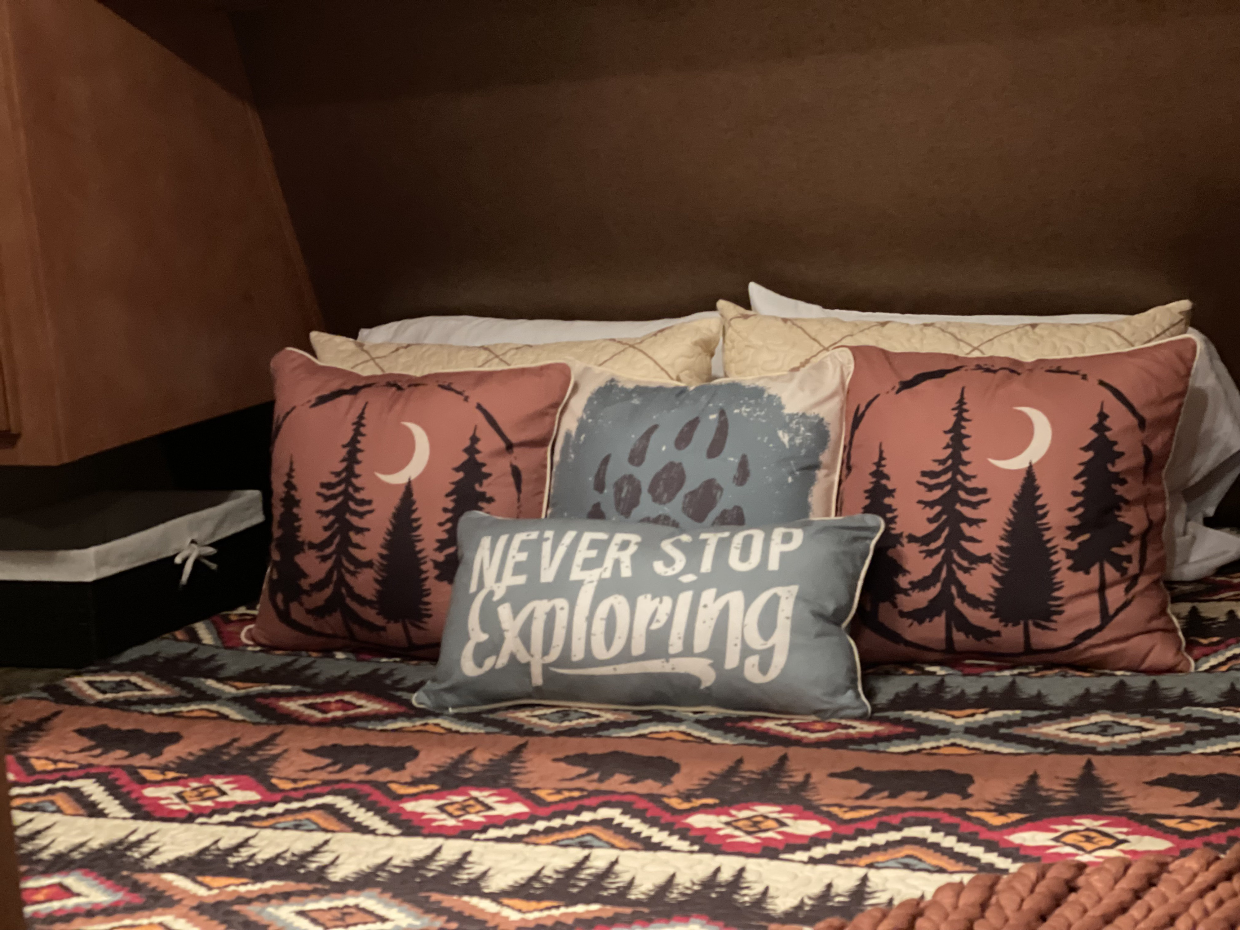 Cozy RV rental bed featuring moon & pine tree pillows, plus a "NEVER STOP EXPLORING" pillow. Rustic bear & tree quilt. Ideal for travel comfort.. Gulf Stream Innsbruck 2014