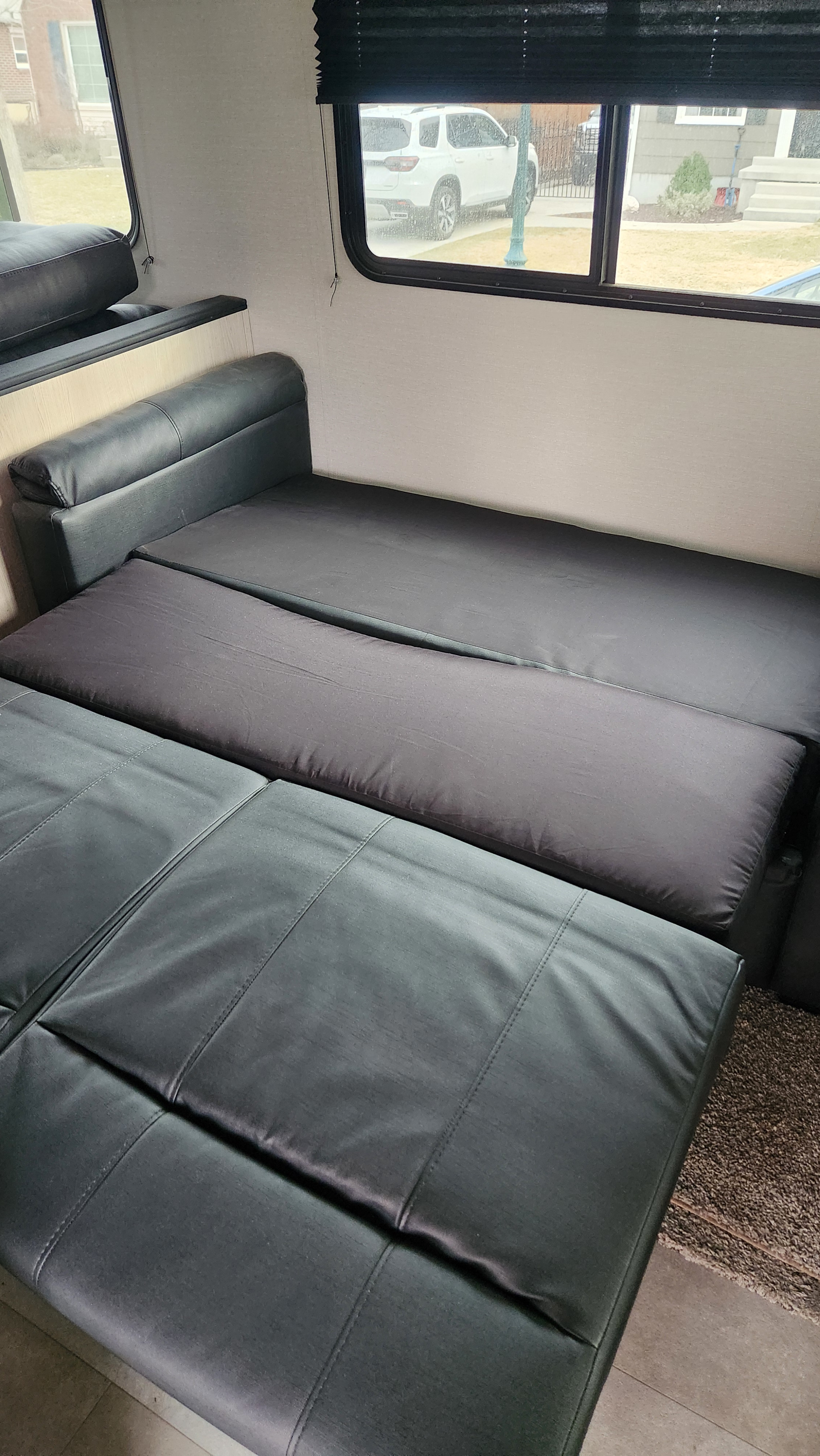 Black convertible sofa/bed inside an RV rental. Sofa is converted for sleeping. Features light walls & windows with outdoor view.. Coachmen Apex 2021