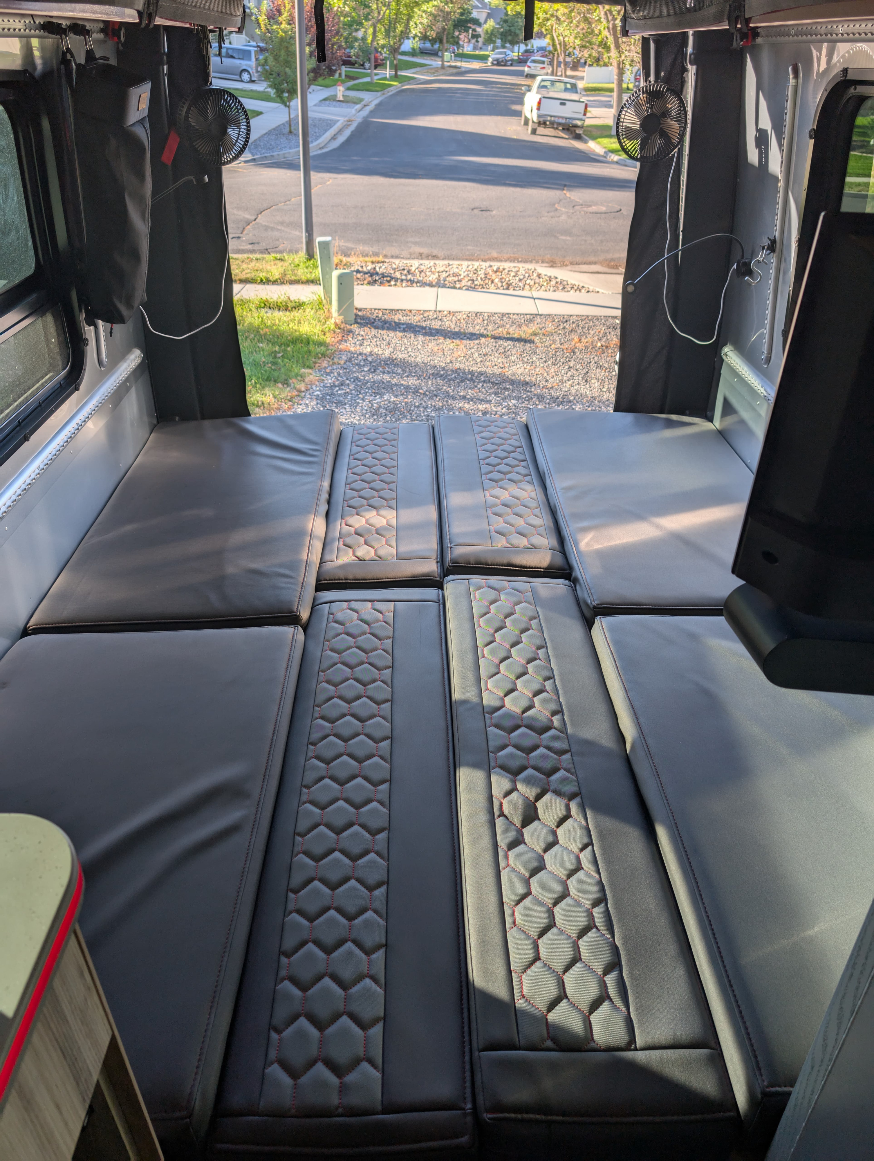 This rv rental features modular black cushions, some with quilted red stitching, arranged as bedding, looking out to a street.. Airstream Interstate 2022