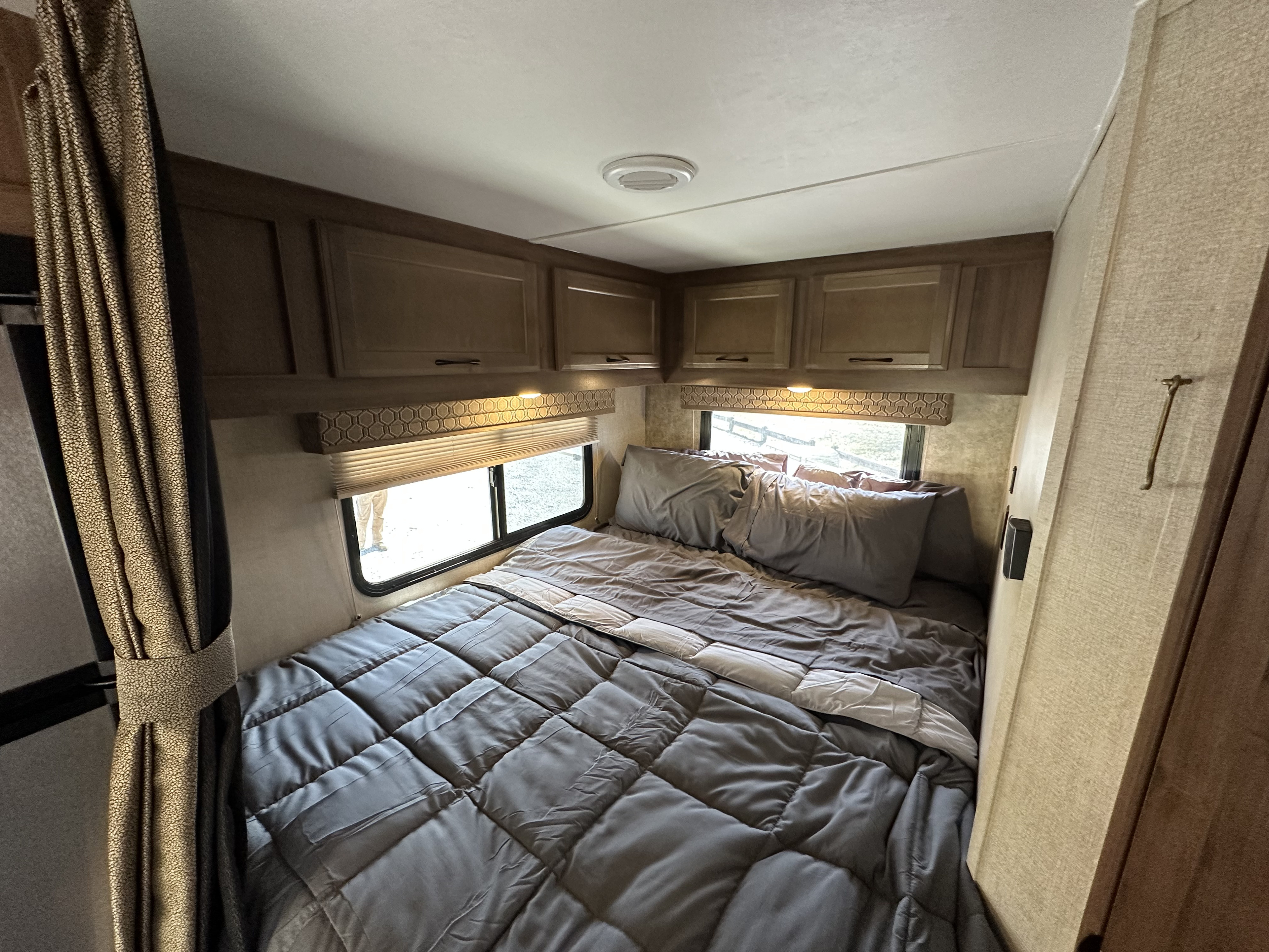 This RV rental bedroom showcases a compact yet cozy bed with grey bedding, overhead cabinets, and a window. Ideal for travel.. Gulf Stream Conquest 2018