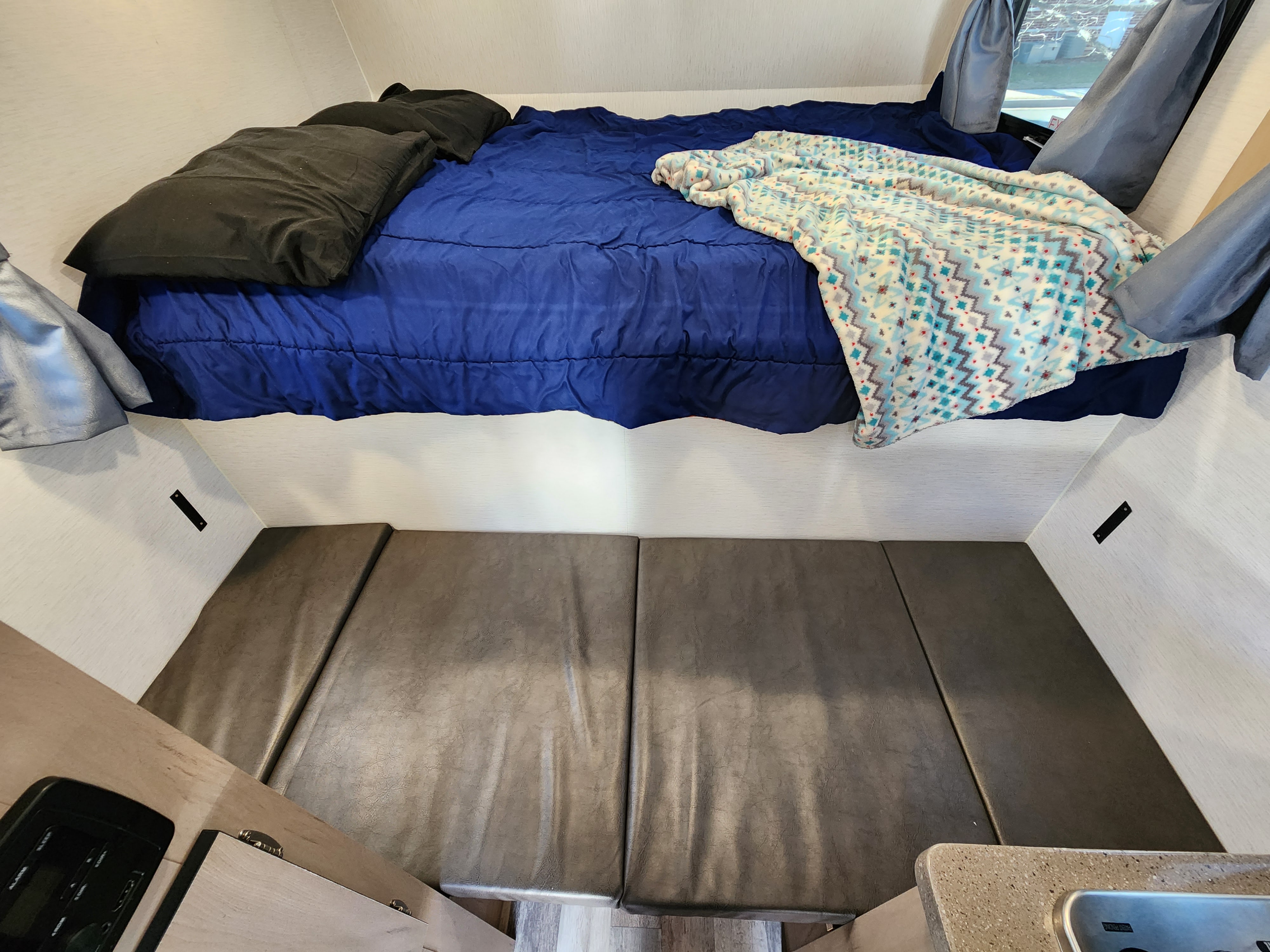 Cozy RV rental interior features a bed with blue comforter & patterned blanket, plus dark grey seating cushions.. Travel Lite Atom 600a 2025