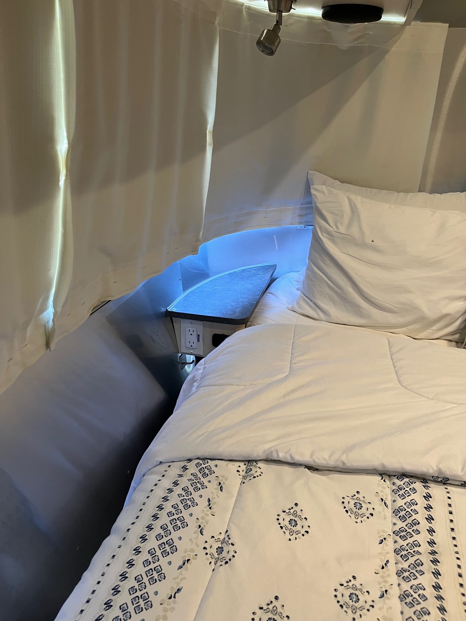Cozy rv rental bedroom: white bedding, patterned duvet, illuminated bedside shelf with outlets, and privacy curtains.. Airstream Flying Cloud 2021