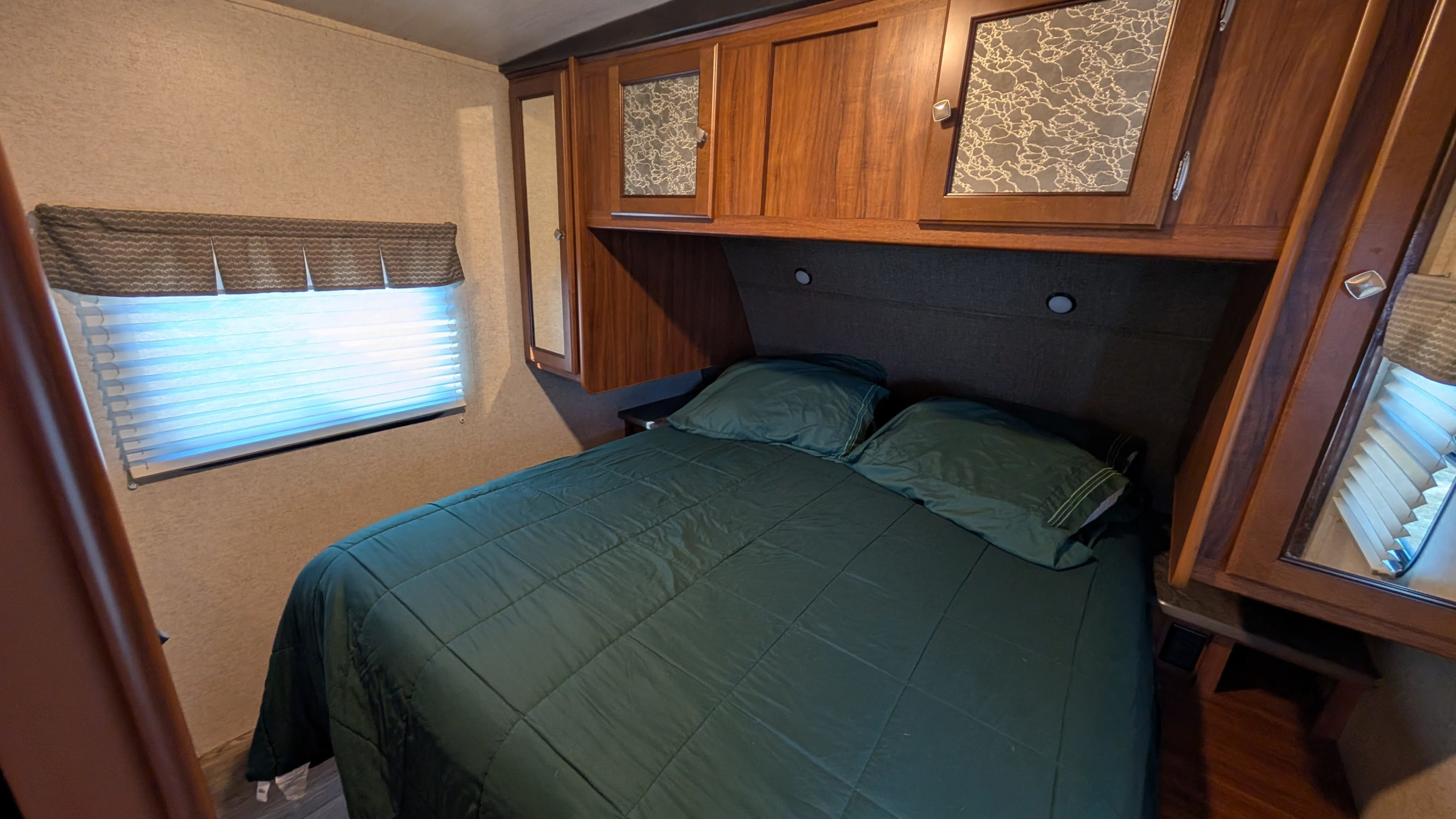 Inside an RV rental: a compact bedroom with a green-quilted bed, overhead storage, and a bright window with blinds.. Heartland RVs Mallard 2018