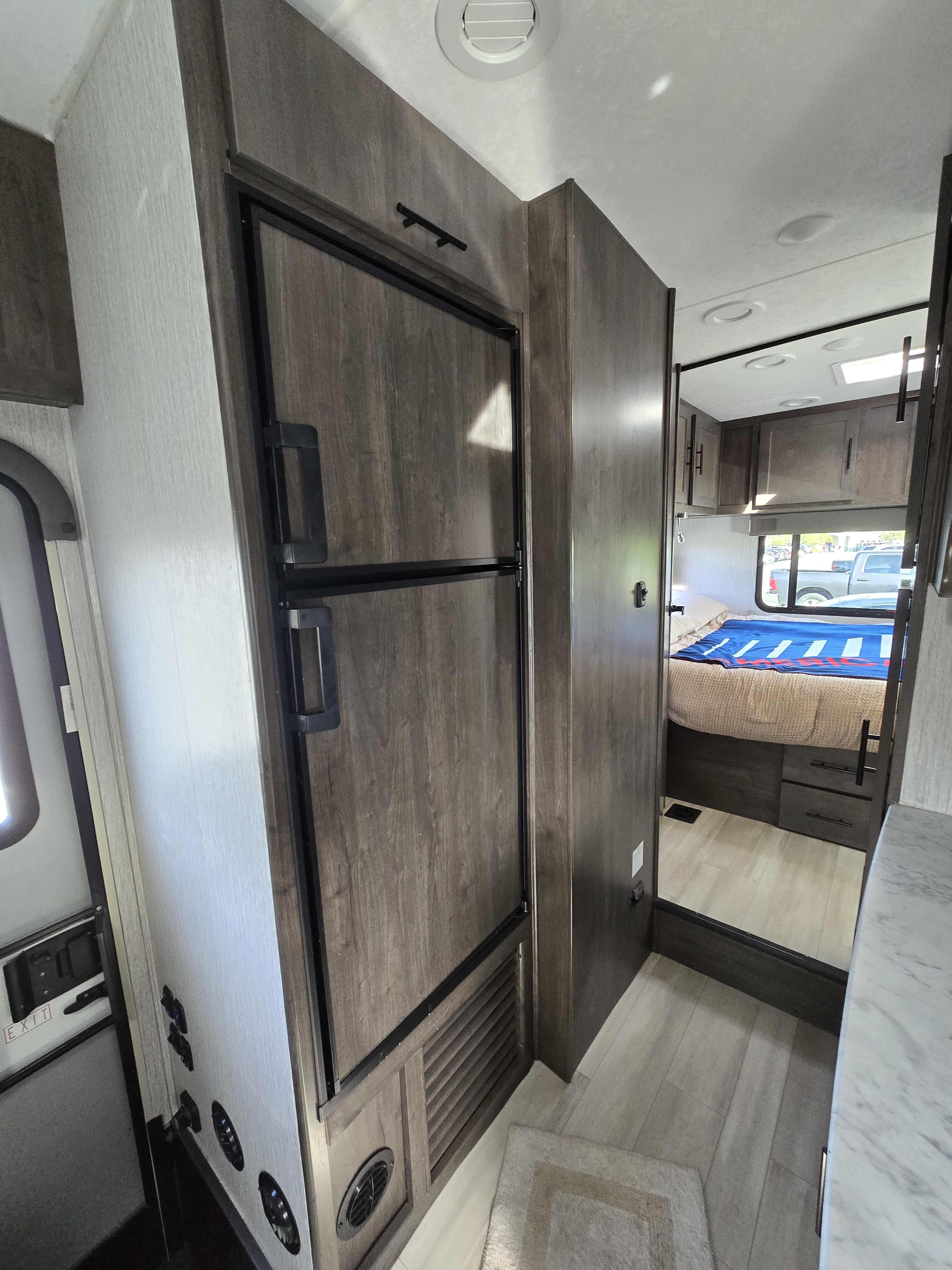 Inside an RV rental, a sleek, dark wood refrigerator is prominent, with the cozy bedroom and light flooring visible beyond.. Coachmen Leprechaun 2023