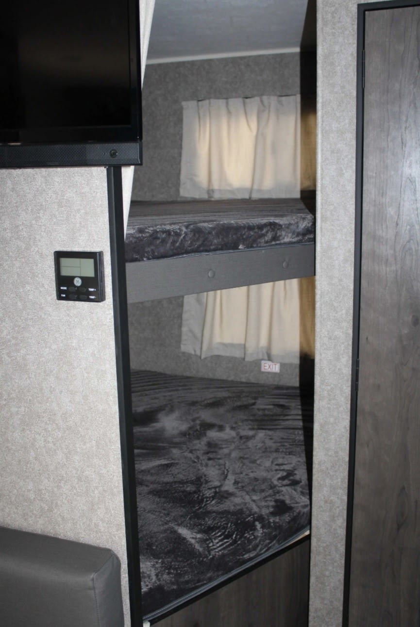 RV rental interior: two bunk beds with mattresses, windows, "EXIT" sign, TV, and digital thermostat shown.. Open Range Open Range 2021