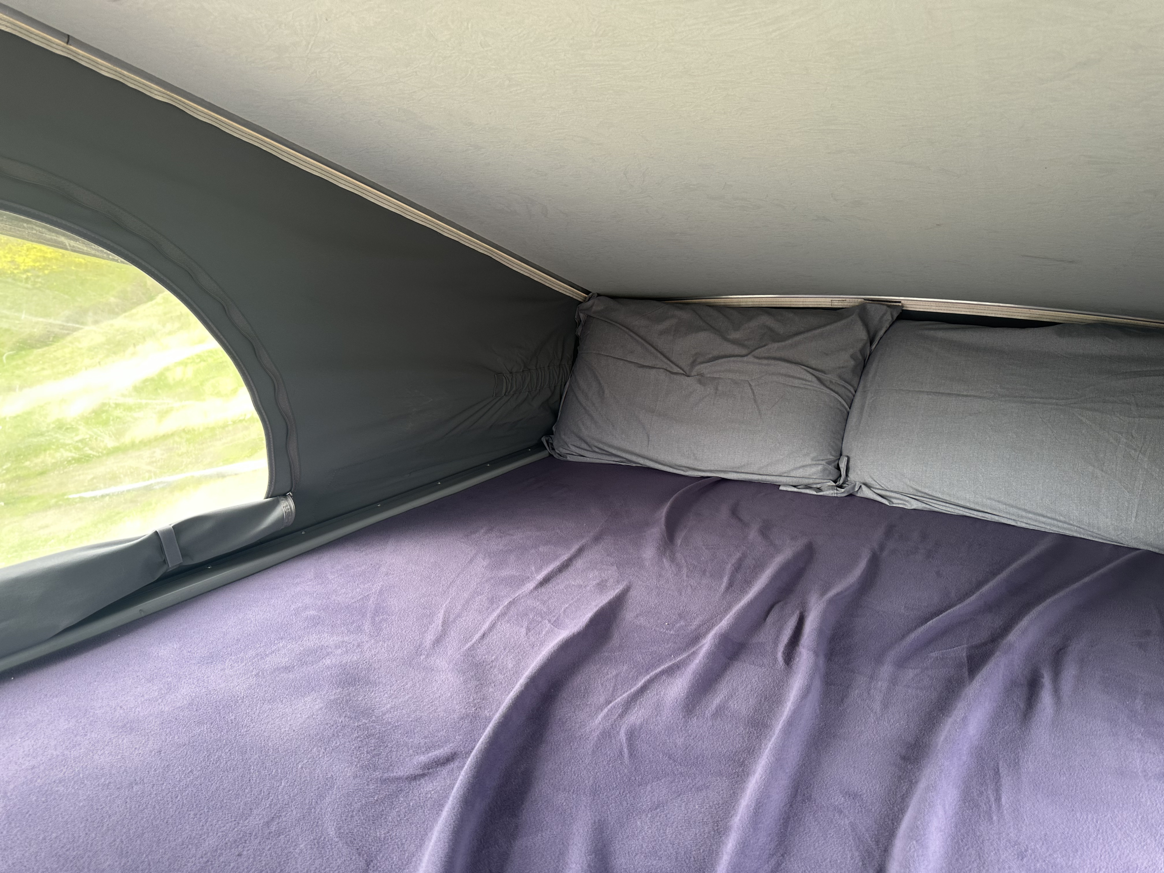 Comfortable sleeping area inside an RV rental's pop-top tent, featuring a purple blanket, grey pillows, and outdoor view.. Winnebago Solis 2023