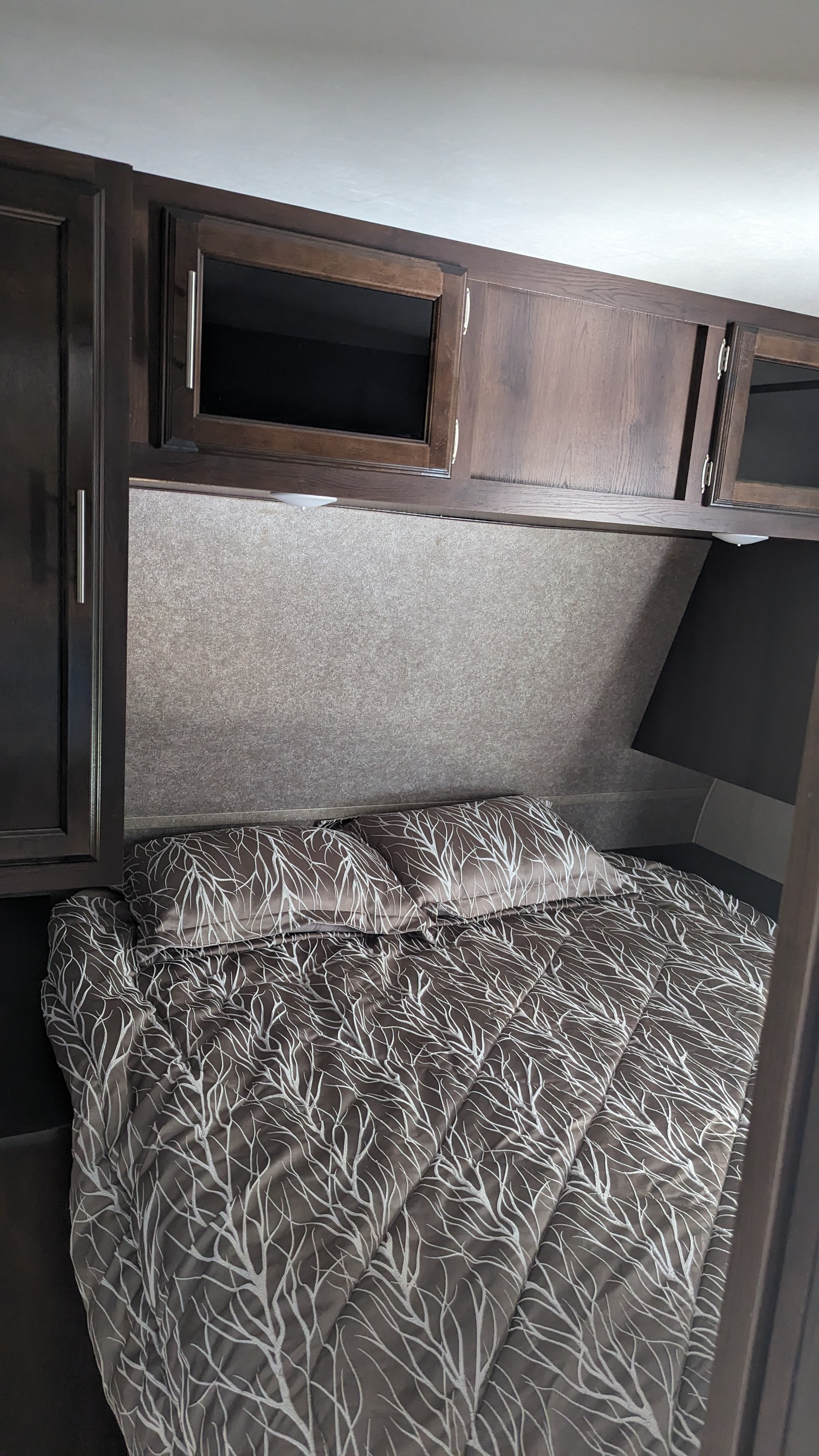 Cozy RV rental bedroom featuring a brown bed with white tree pattern, dark wood overhead cabinets, and textured headboard.. Jayco Jay Flight 2019