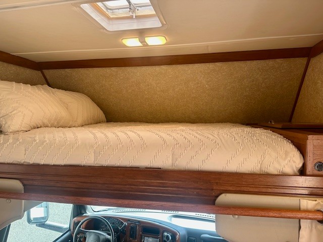 Inside an RV rental: a bright, cozy overcab bed with white bedding, ideal for travel. Dashboard visible below.. Four Winds Four Winds Motorhome 2006