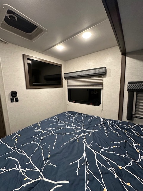 Inside a modern RV rental: a comfortable bedroom with a patterned duvet, TV, and adjustable window blinds.. Thor Motor Coach Four Winds 22B 2025