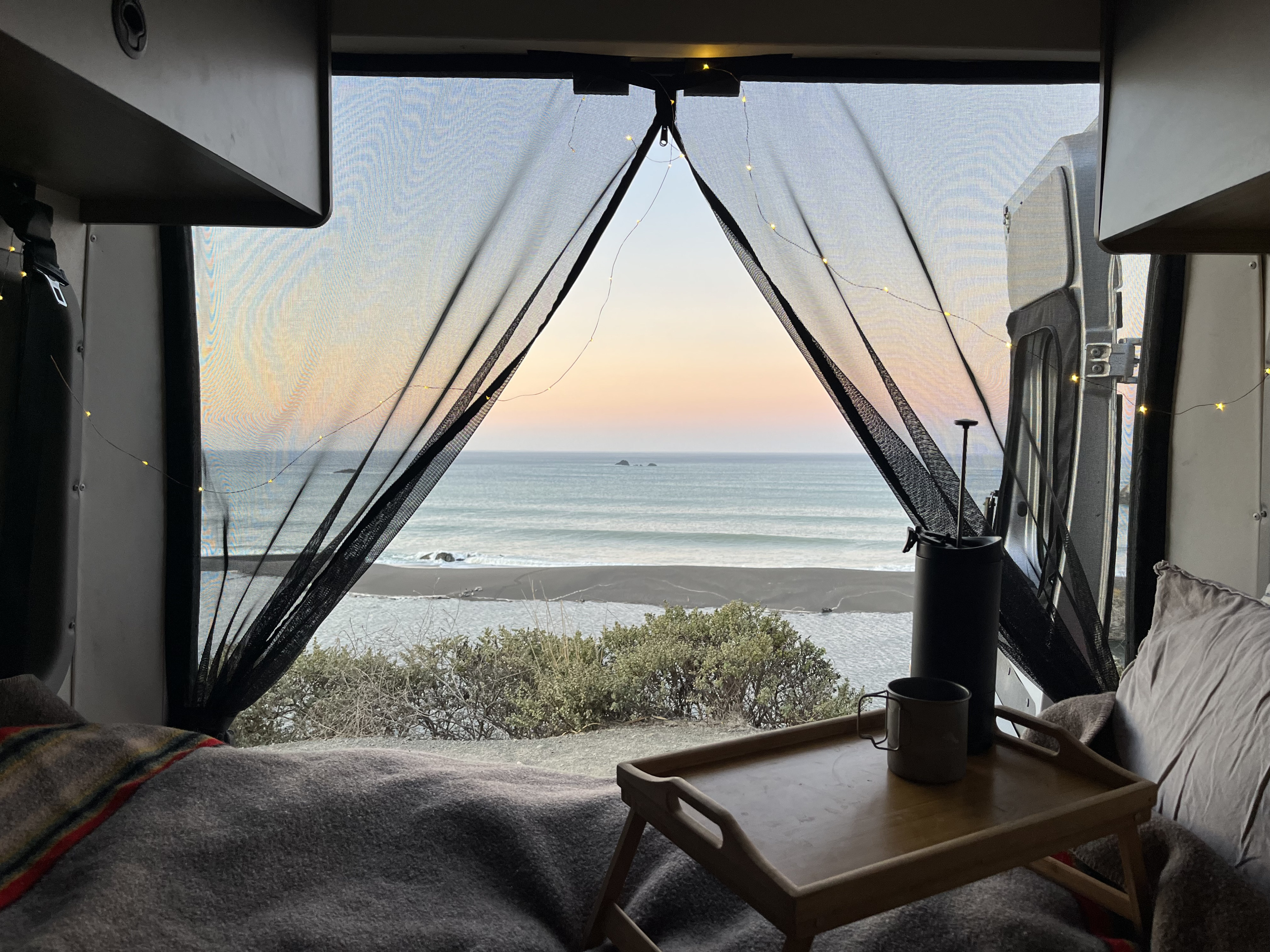 Inside an RV rental: a tranquil bedroom view of the ocean, beach, and sunset with string lights.. Winnebago Solis 2022