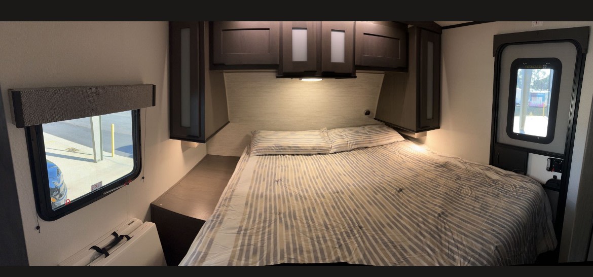 An RV rental's cozy bedroom, showcasing a striped bed, dark overhead cabinets, a bedside shelf, and bright windows.. Cruiser RV MPG Ultra Lite 2022