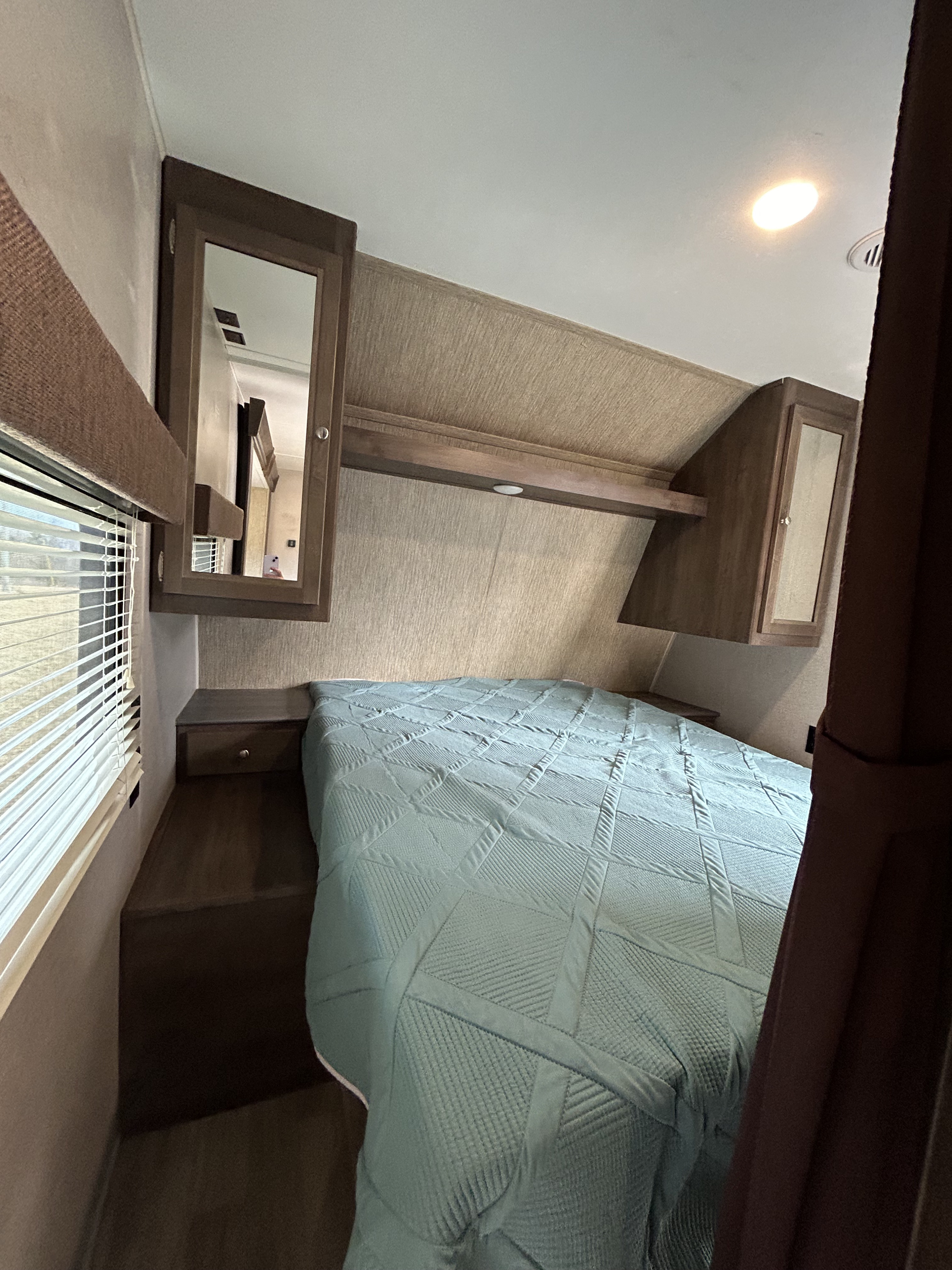 Inside an RV rental bedroom: a cozy bed with a teal quilt, window, and ample overhead storage. Perfect for travel comfort.. Dutchmen Coleman Lantern 2019