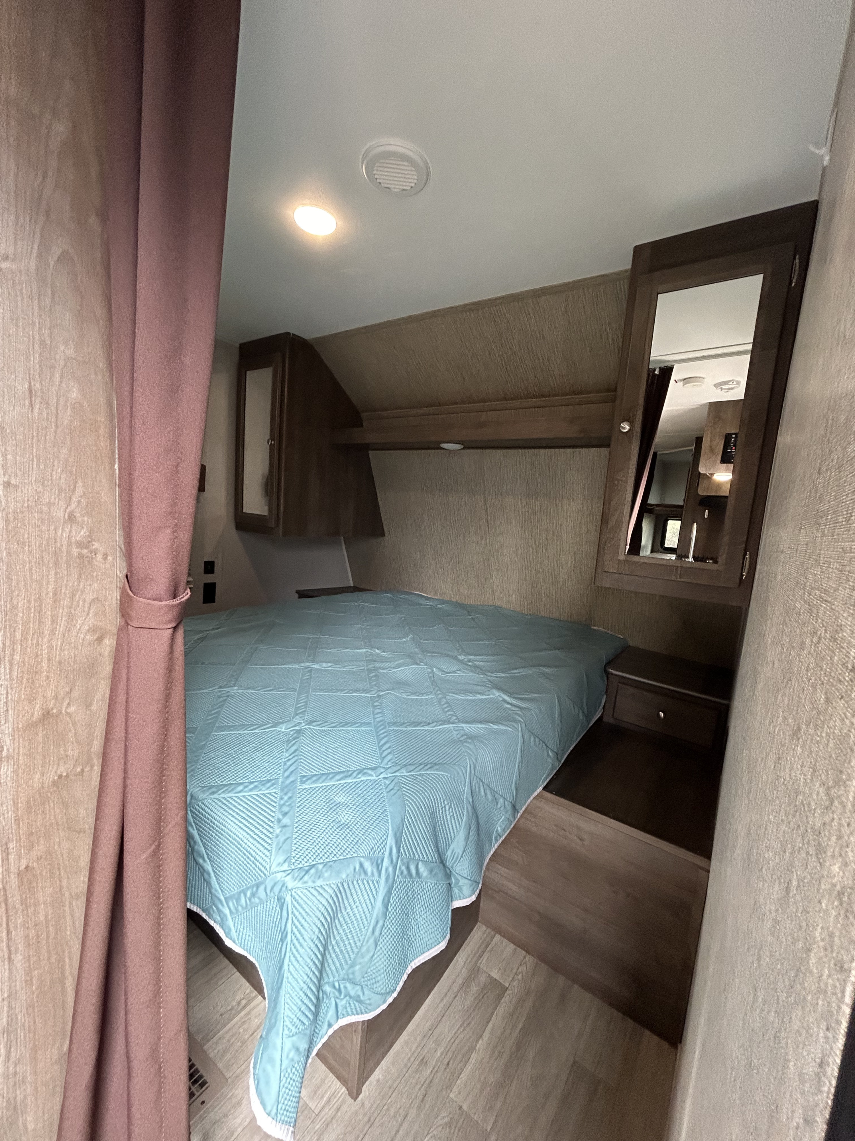 Inside an RV rental: a cozy bedroom with a blue quilted bed, built-in storage, a mirror, and light wood interior.. Dutchmen Coleman Lantern 2019