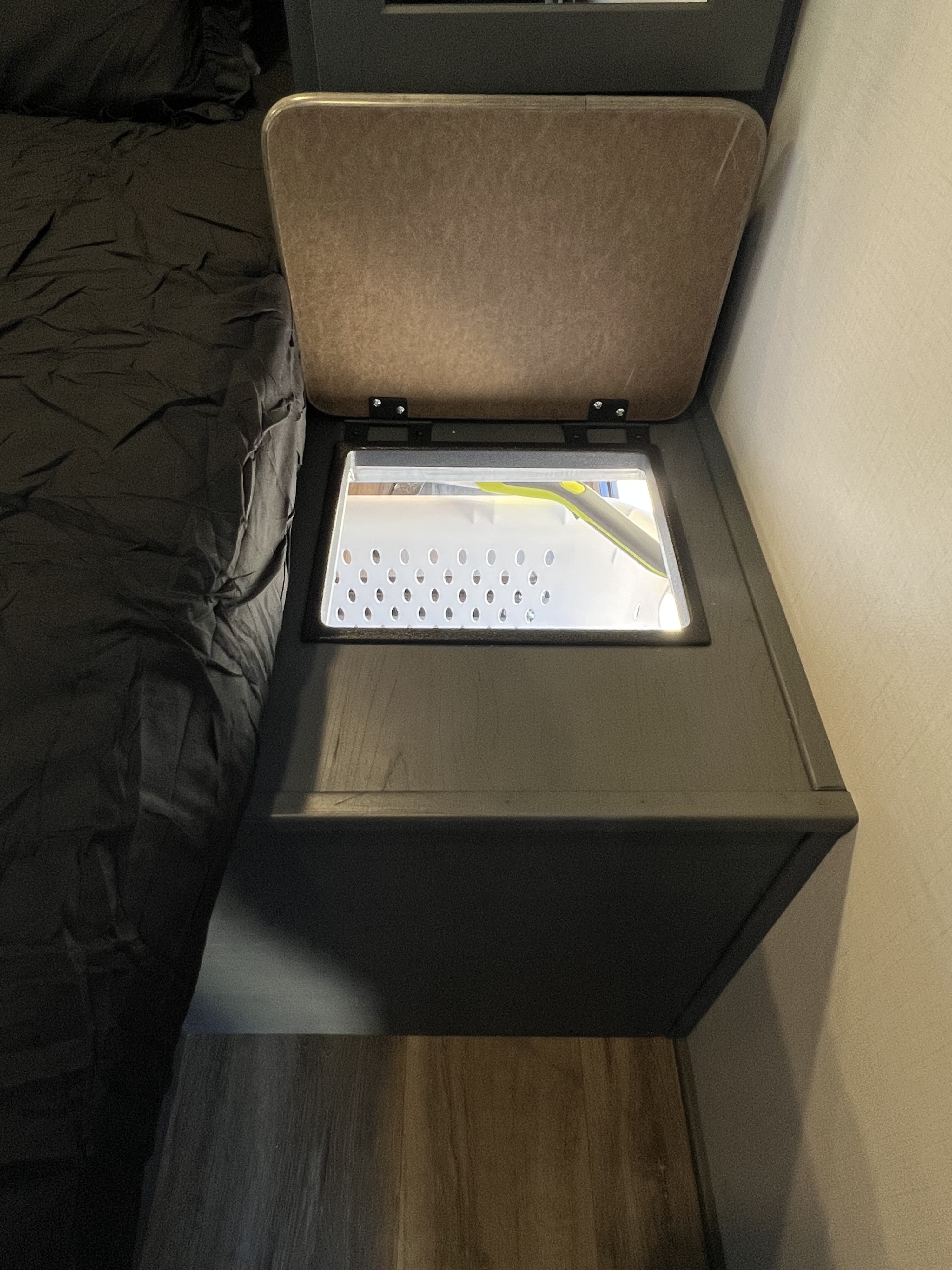 Illuminated bedside storage in an RV rental, showing a perforated bin and bright green tool next to dark bedding.. Keystone RV Bullet Ultra Lite 2024