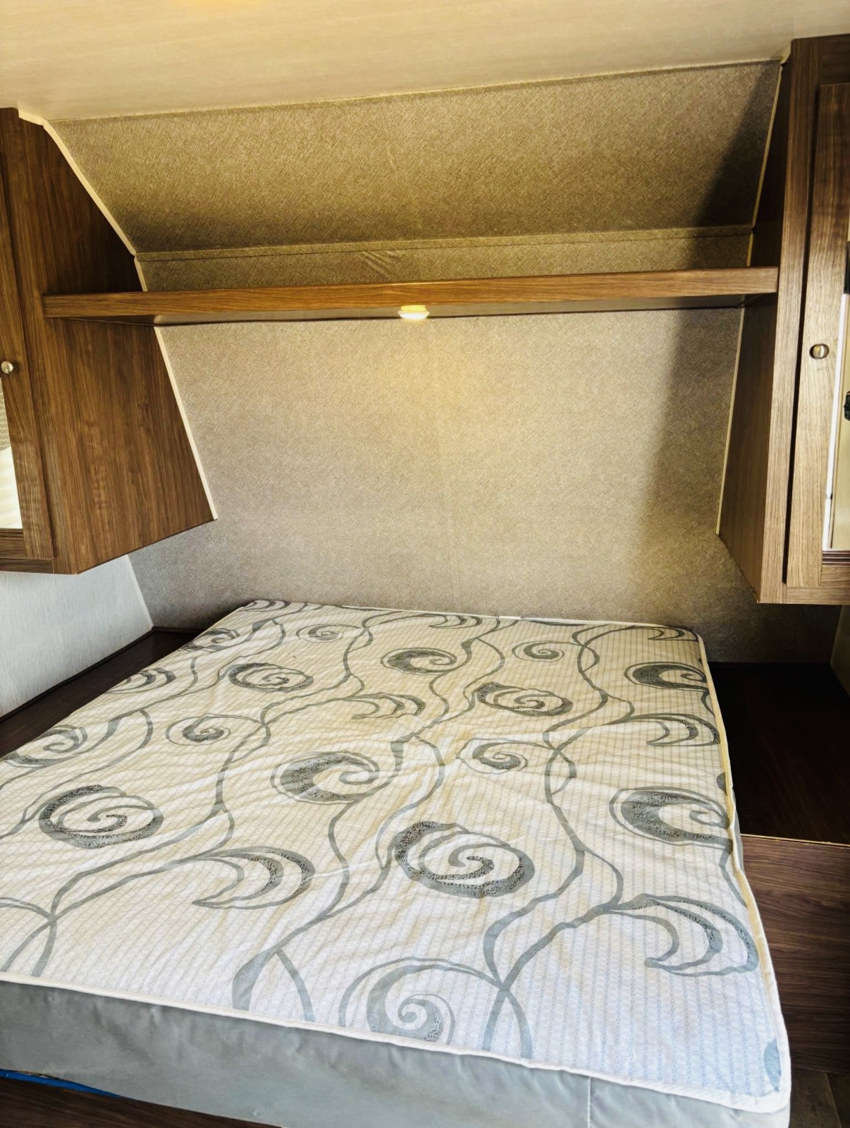 Cozy RV rental bedroom featuring a bed with a patterned mattress, wood cabinets, and overhead shelving with a light.. Heartland RVs Trail Runner SLE 2018