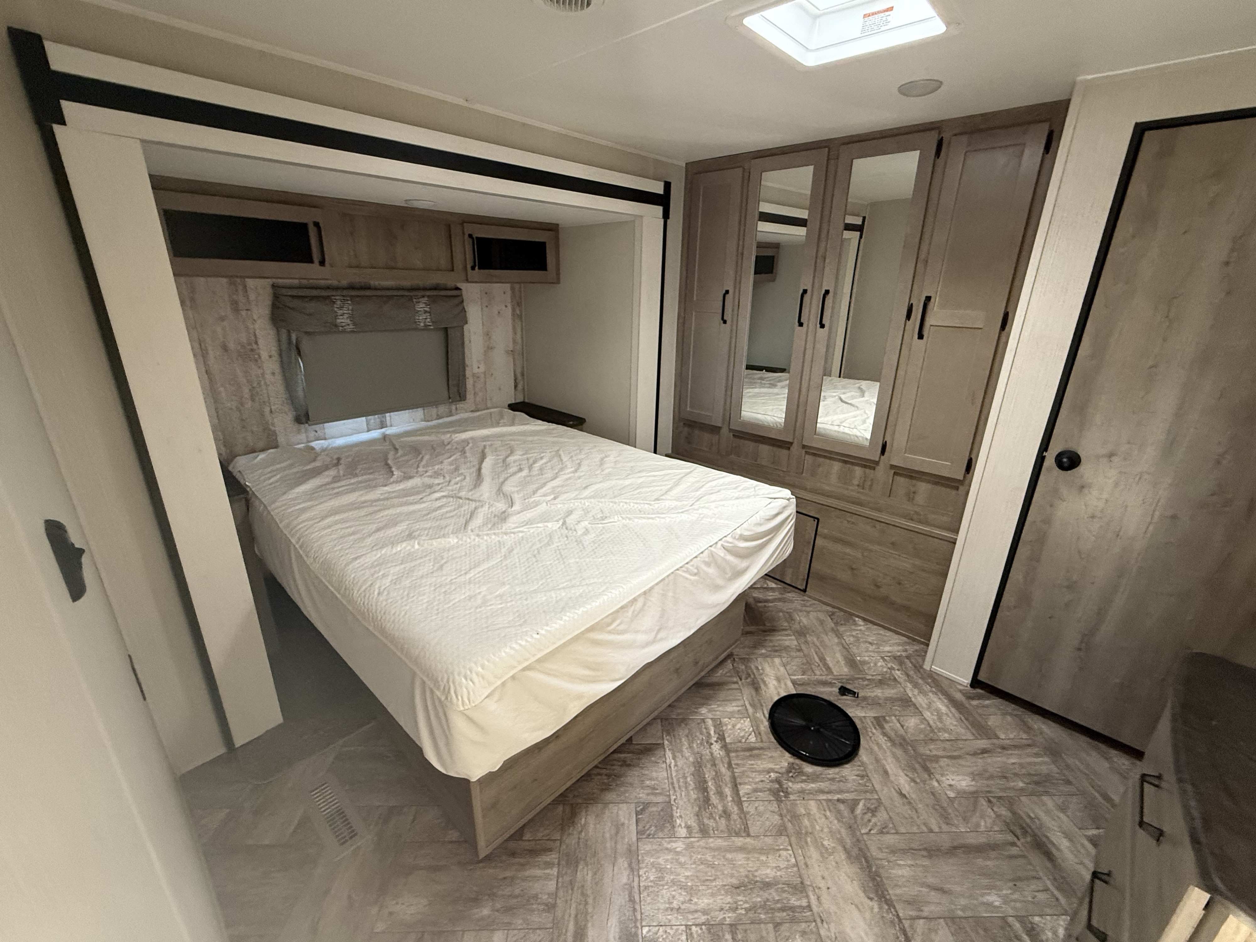 Modern RV rental bedroom with a neatly made bed, overhead cabinets, and a mirrored wardrobe. Wood-look flooring and skylight.. Palomino Puma 2023