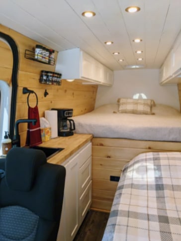 This bright **RV rental** interior showcases a cozy bed, compact kitchen, and overhead storage in a wood-paneled van.. Sprinter Mercedes 2018