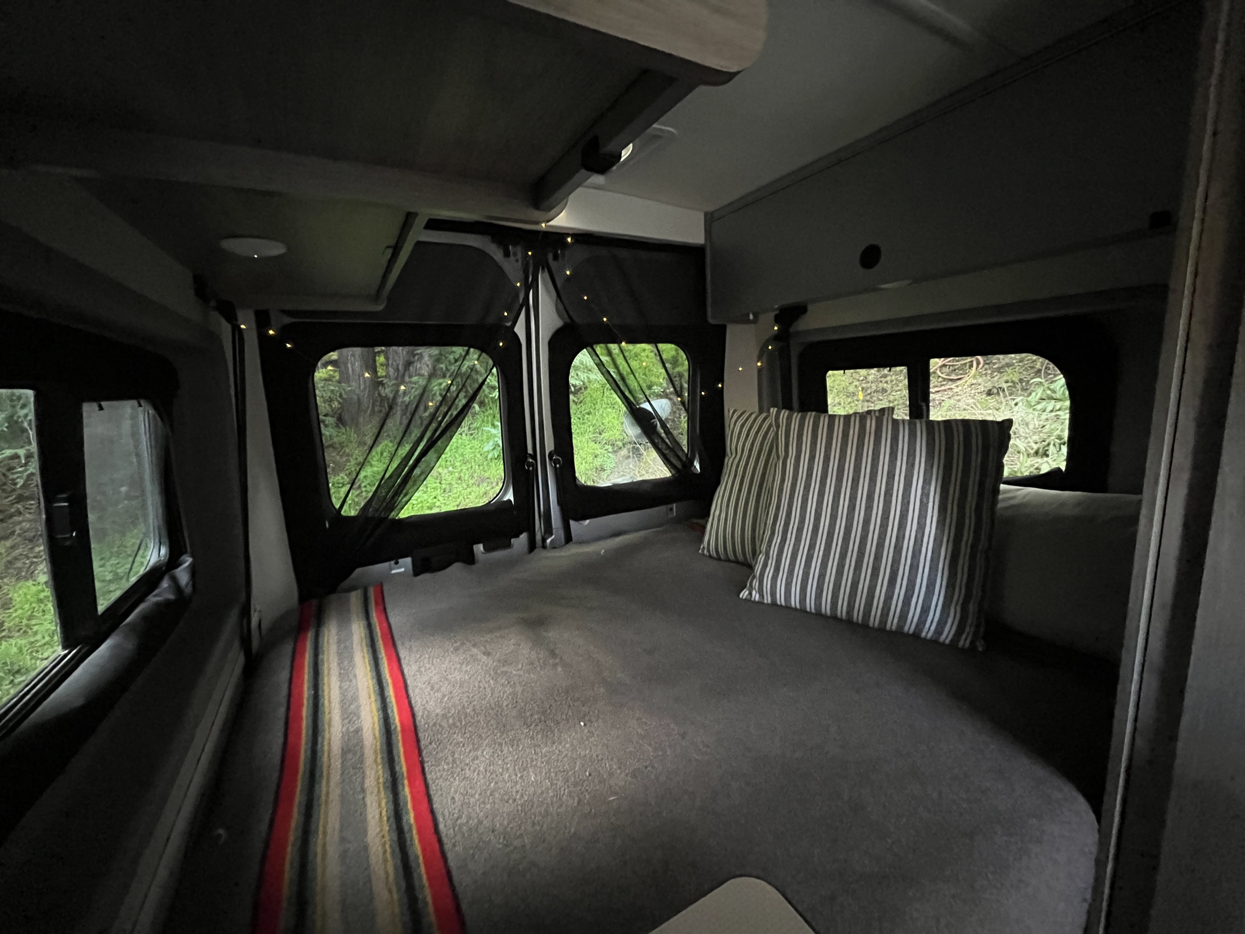 Relax in this cozy RV rental's camper van bedroom. Features a comfortable bed with striped pillows & natural views with string lights.. Winnebago Solis 2022