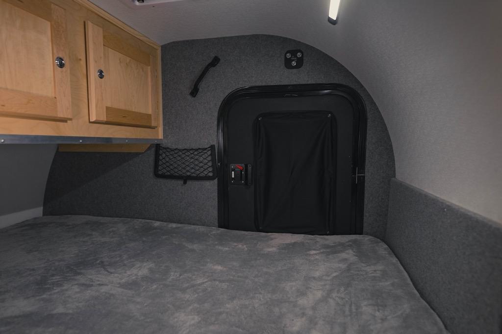 Cozy compact RV rental interior with a comfy bed, wood cabinets, storage net, and an access door.. Sunnyside Boony Stomper 2024