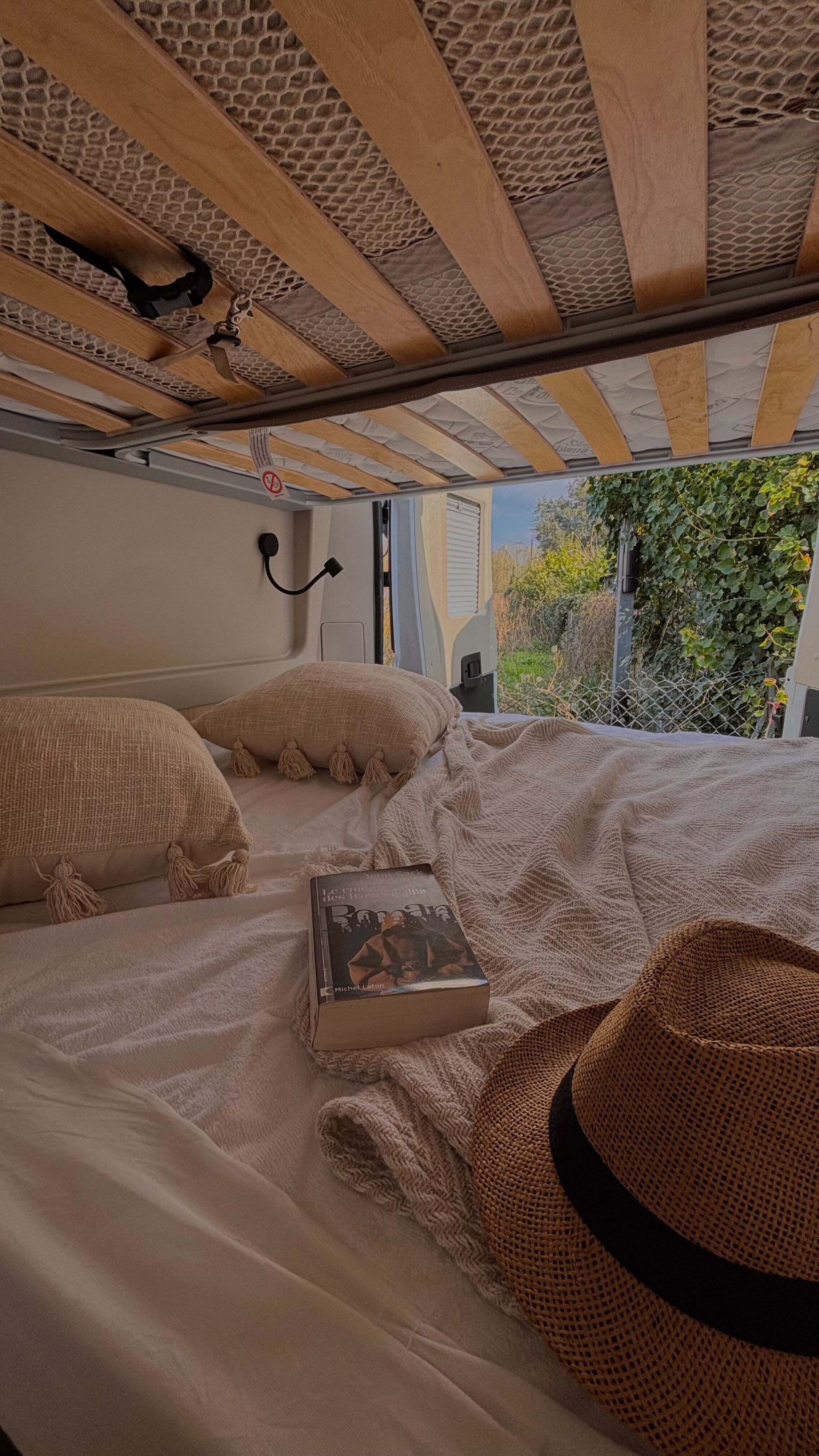 Cozy RV rental bedroom scene: bed with pillows, blanket, book, straw hat, and open back doors to a lush green outdoor view.. Chausson V 594  MAX FIRST LINE 2024