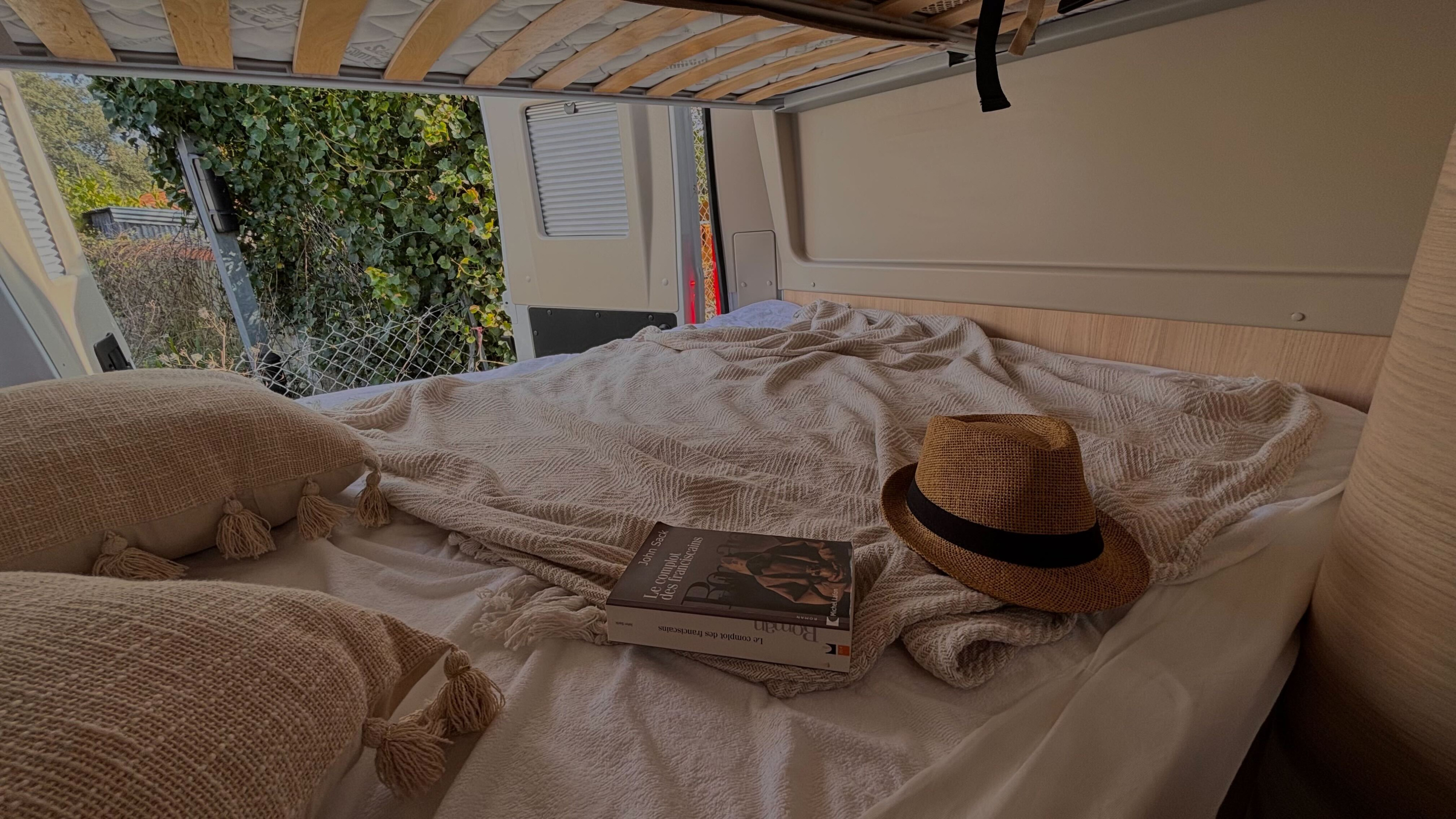 Cozy RV rental bed with textured pillows, blanket, book, and straw hat. Open doors reveal lush greenery.. Chausson V 594  MAX FIRST LINE 2024