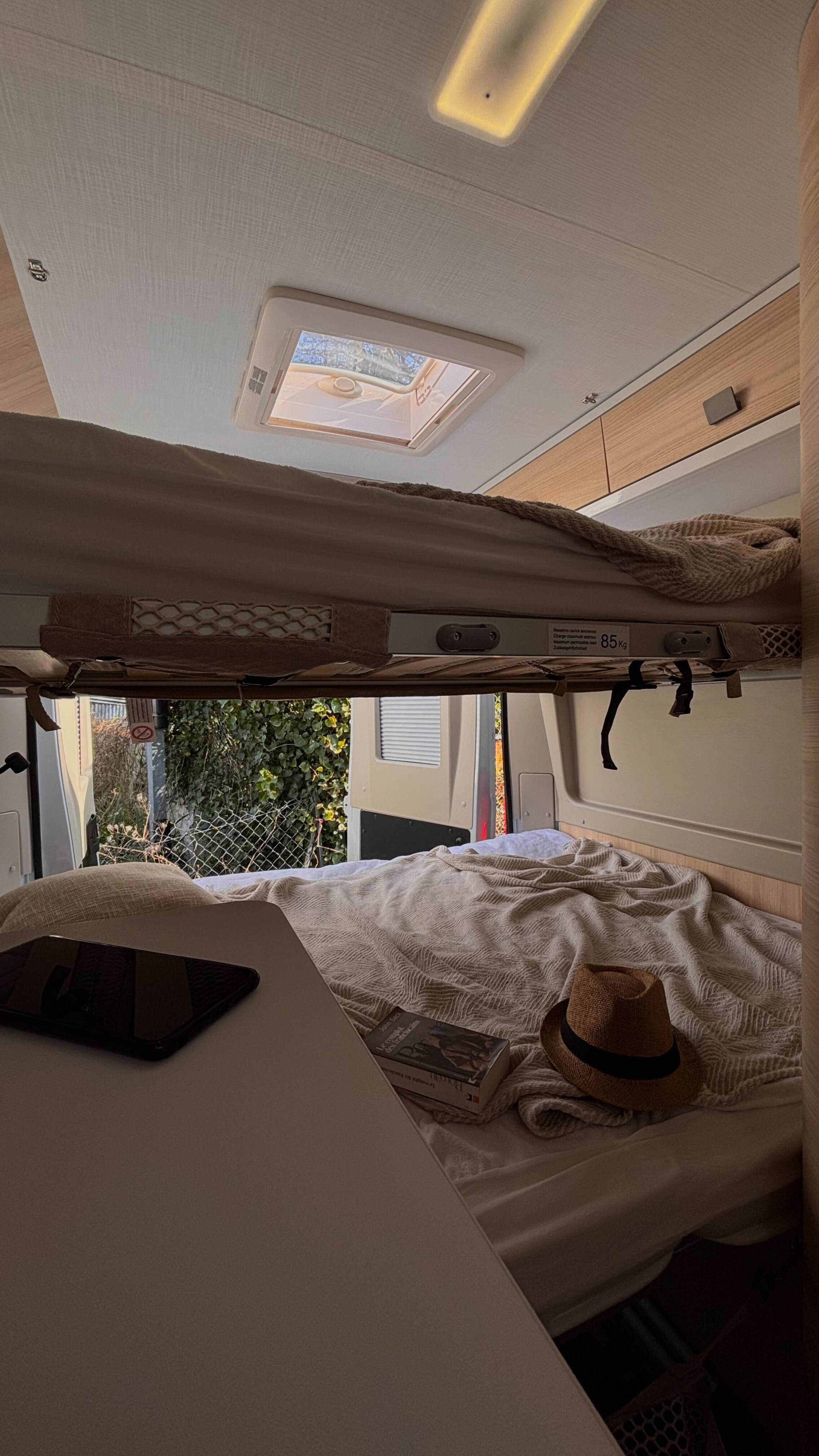 Inside a comfy rv rental, featuring bunk beds, a bright skylight, and a lower bed with a book, hat, and device on a table.. Chausson V 594 MAX FIRST LINE 2024