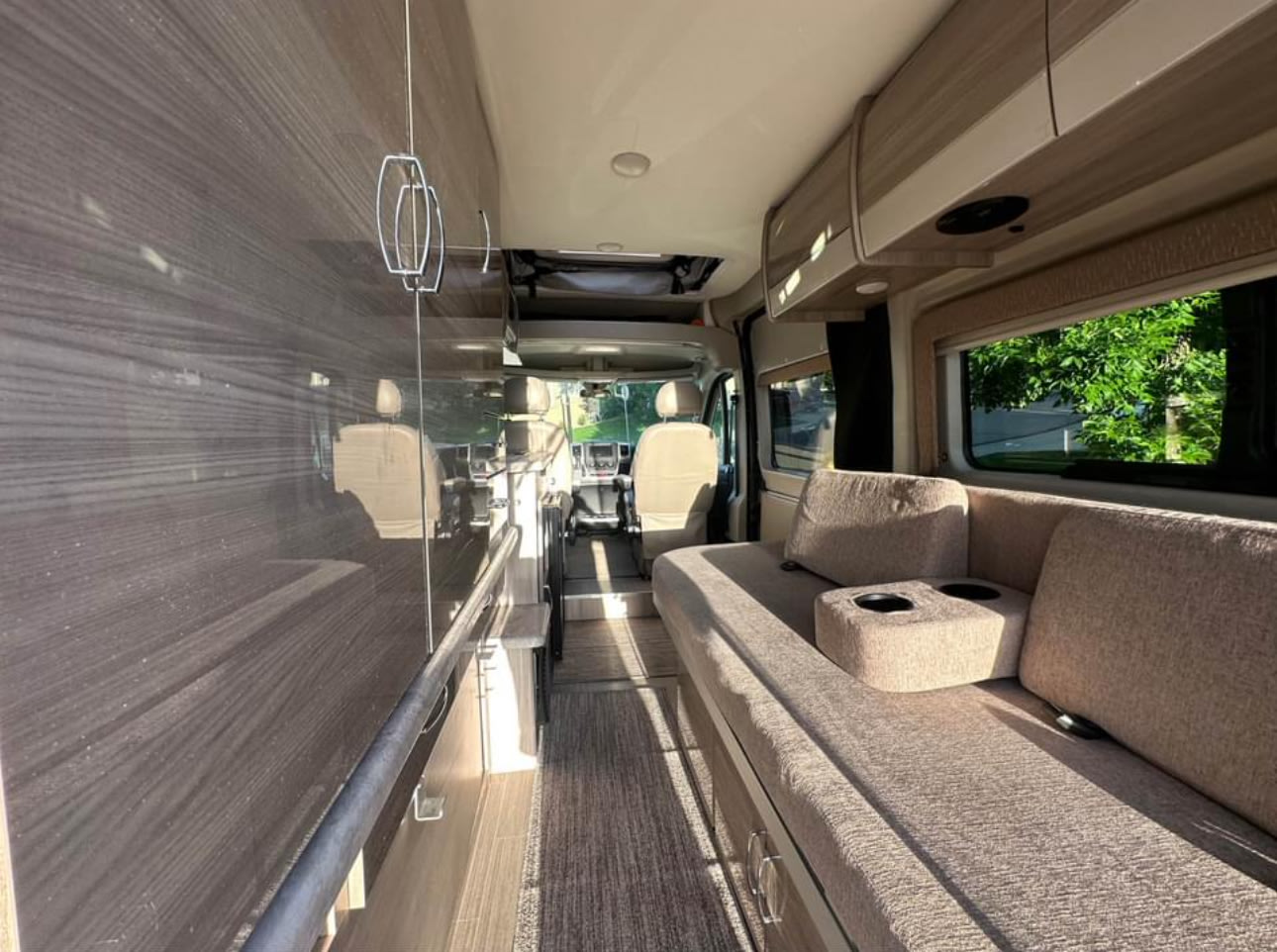 Inside this modern RV rental: a cozy living space with a long couch, overhead cabinets, and bright views.. Thor Motor Coach 18T 2022
