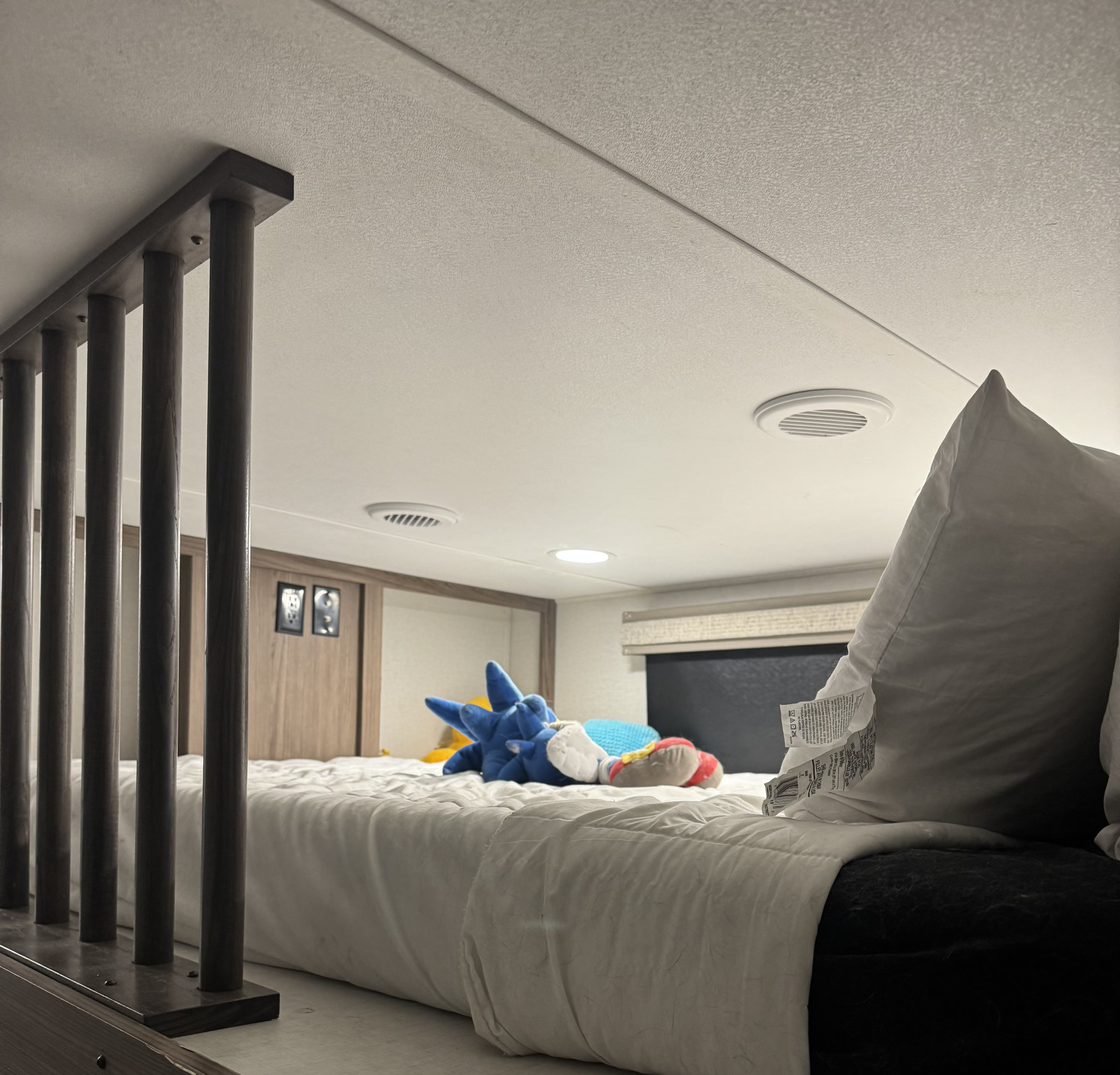 Inside an RV rental: a comfortable loft bed with a white comforter, Sonic plushie, dark wood railing, and ceiling vents.. Alliance Paradigm 2023