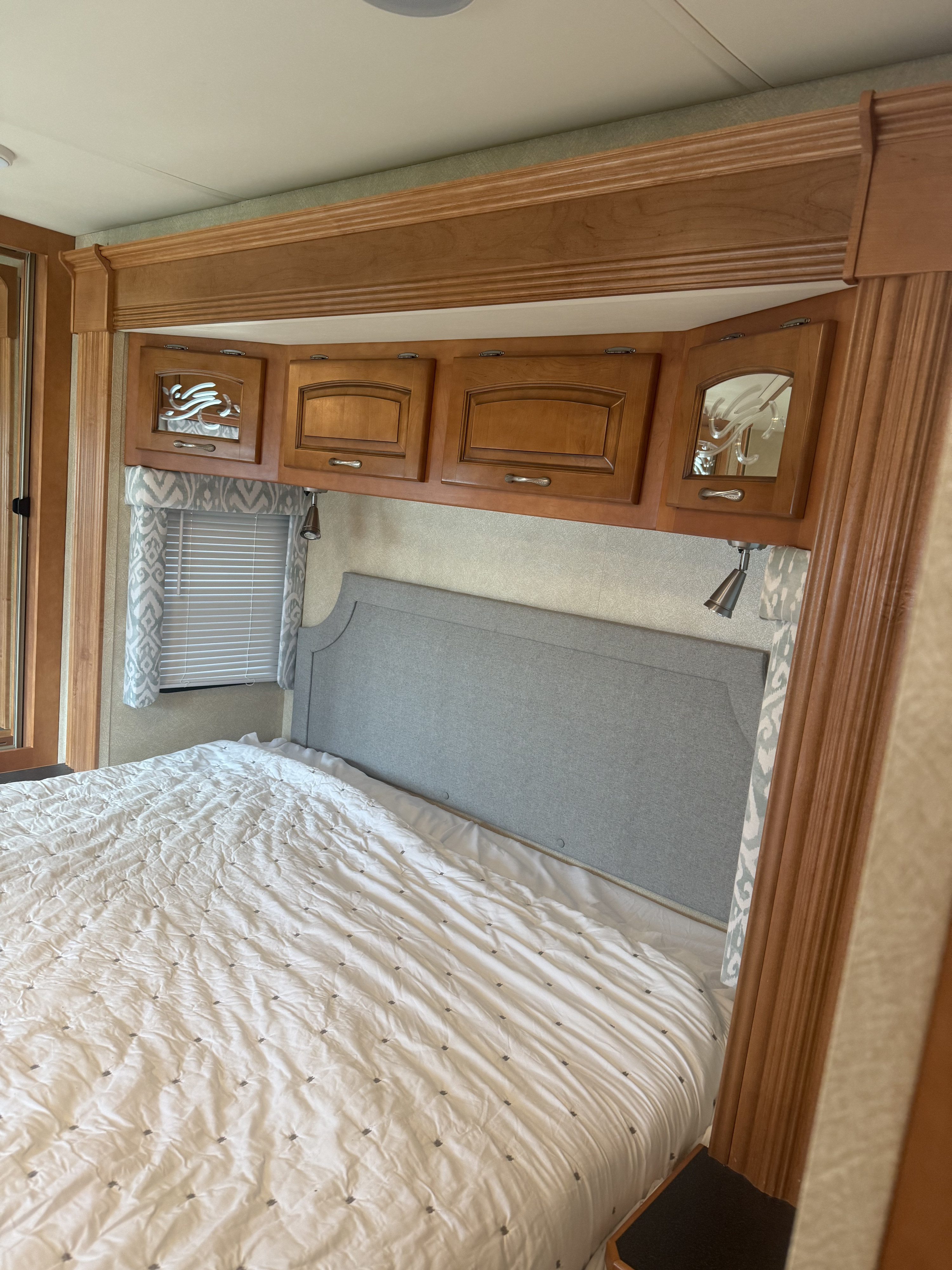 RV rental bedroom with a cozy bed, gray headboard, wooden overhead cabinets, and a window with blinds and valances.. Kountry Star Kountry Star Motorhome 2008