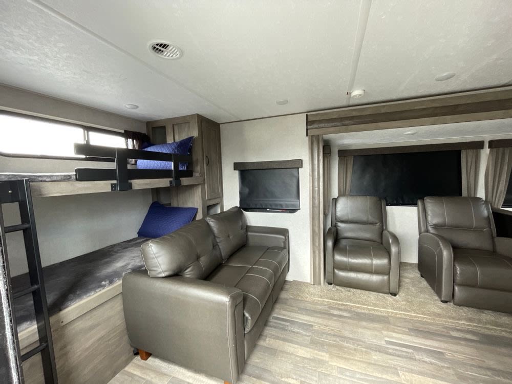 This RV rental's interior shows a living space with a gray sofa, two recliners, and bunk beds with blue pillows.. Forest River Vibe 2021