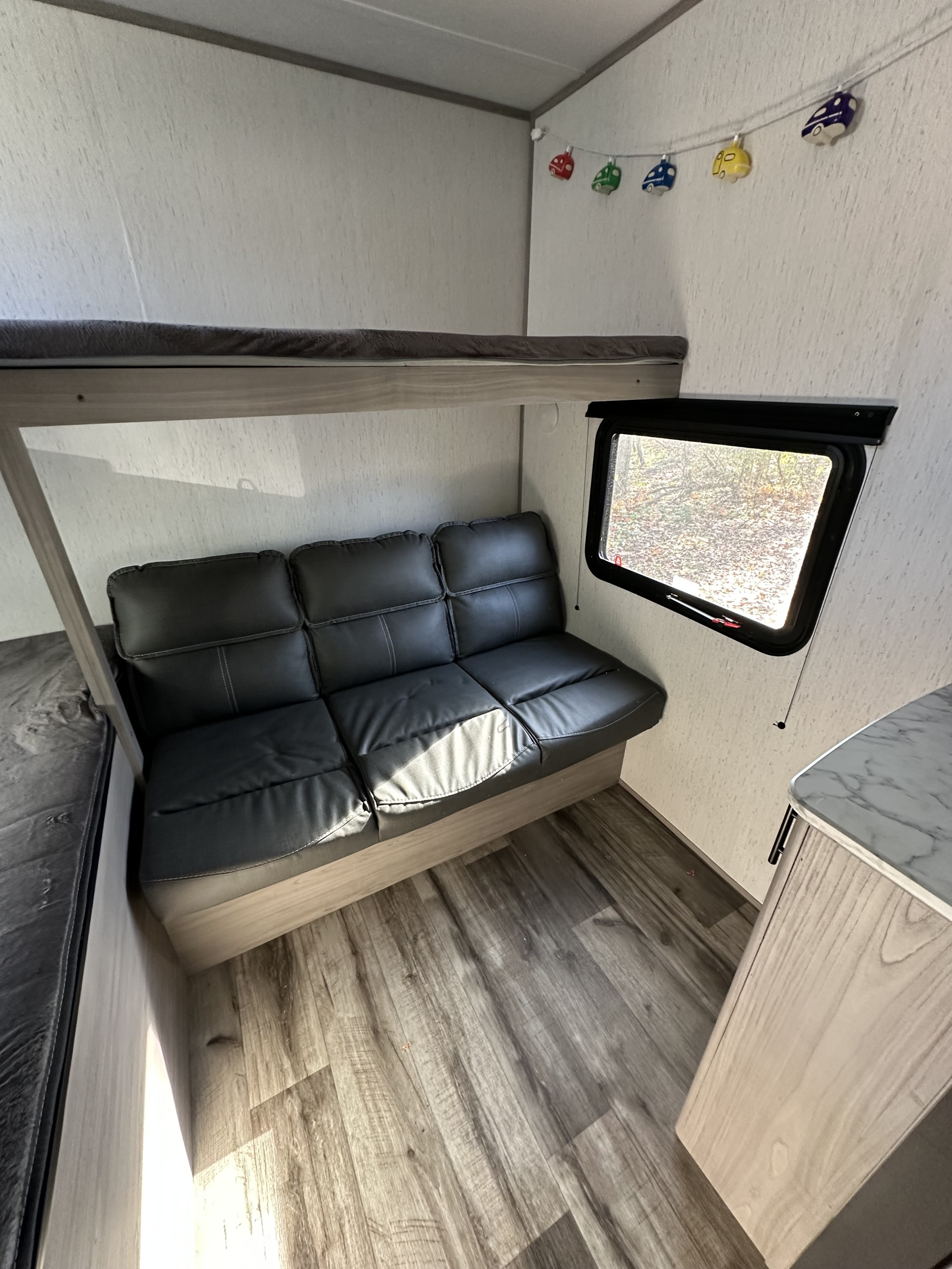 Inside an RV rental: a black couch under a top bunk, a window view, and fun car string lights.. Dutchmen Coleman Lantern 2022