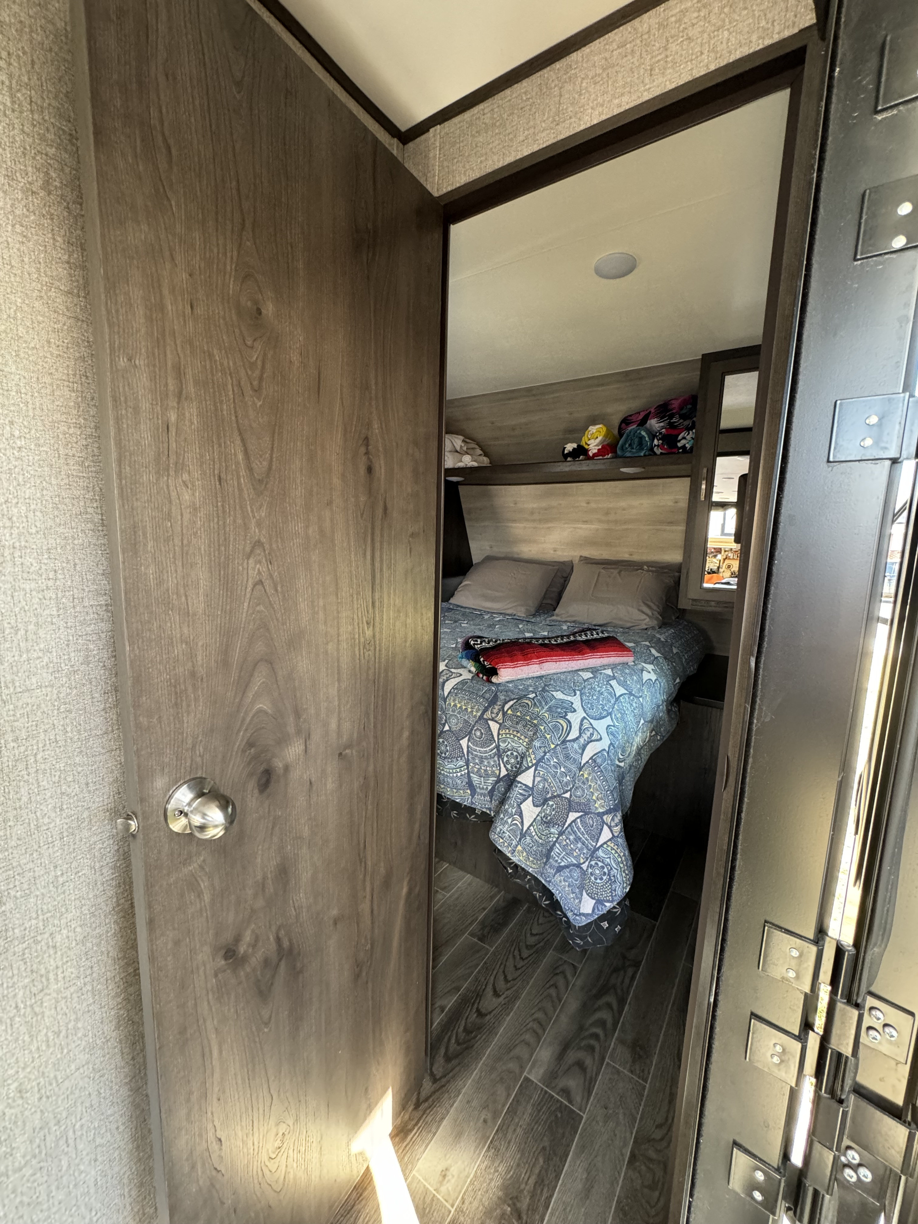 Inside an RV rental: A cozy bedroom with patterned bedding and overhead shelf is visible through a partially open wooden door.. Jayco Jay Flight 2020