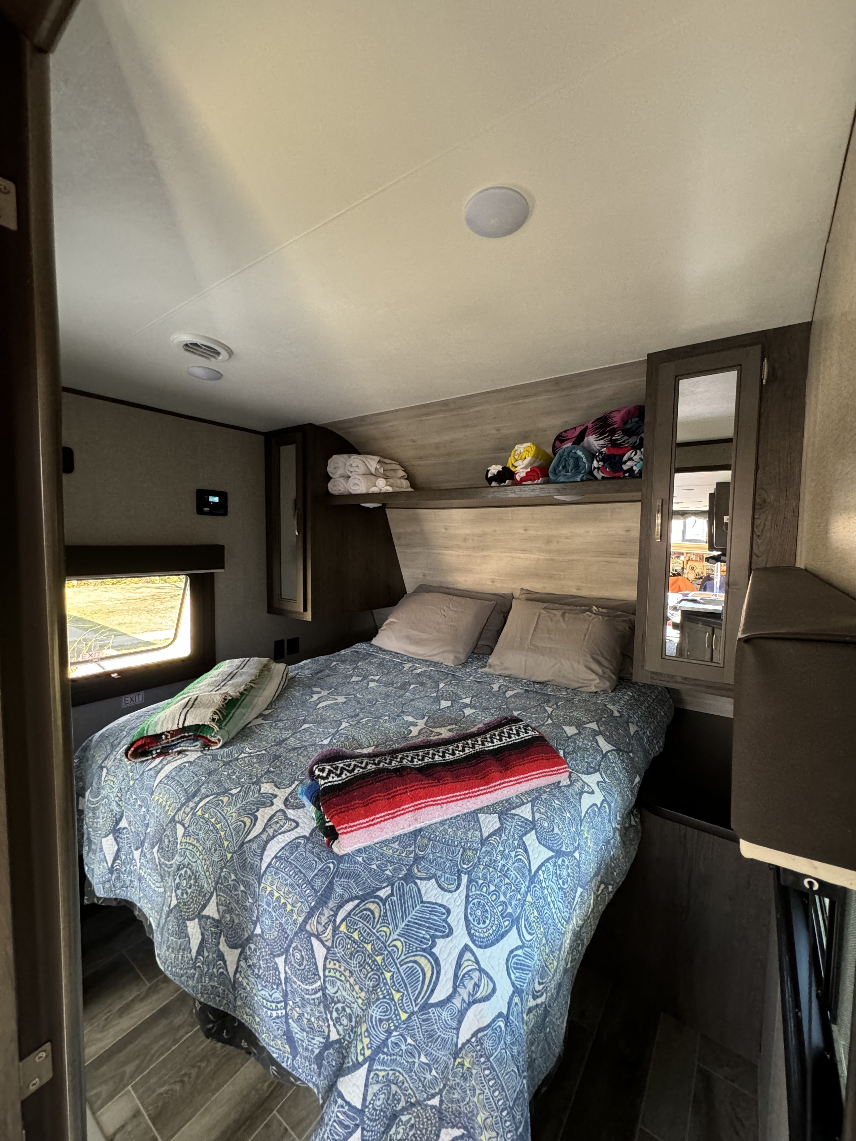 An RV rental bedroom features a patterned bed with throws, built-in storage, and a window looking out onto greenery.. Jayco Jay Flight 2020