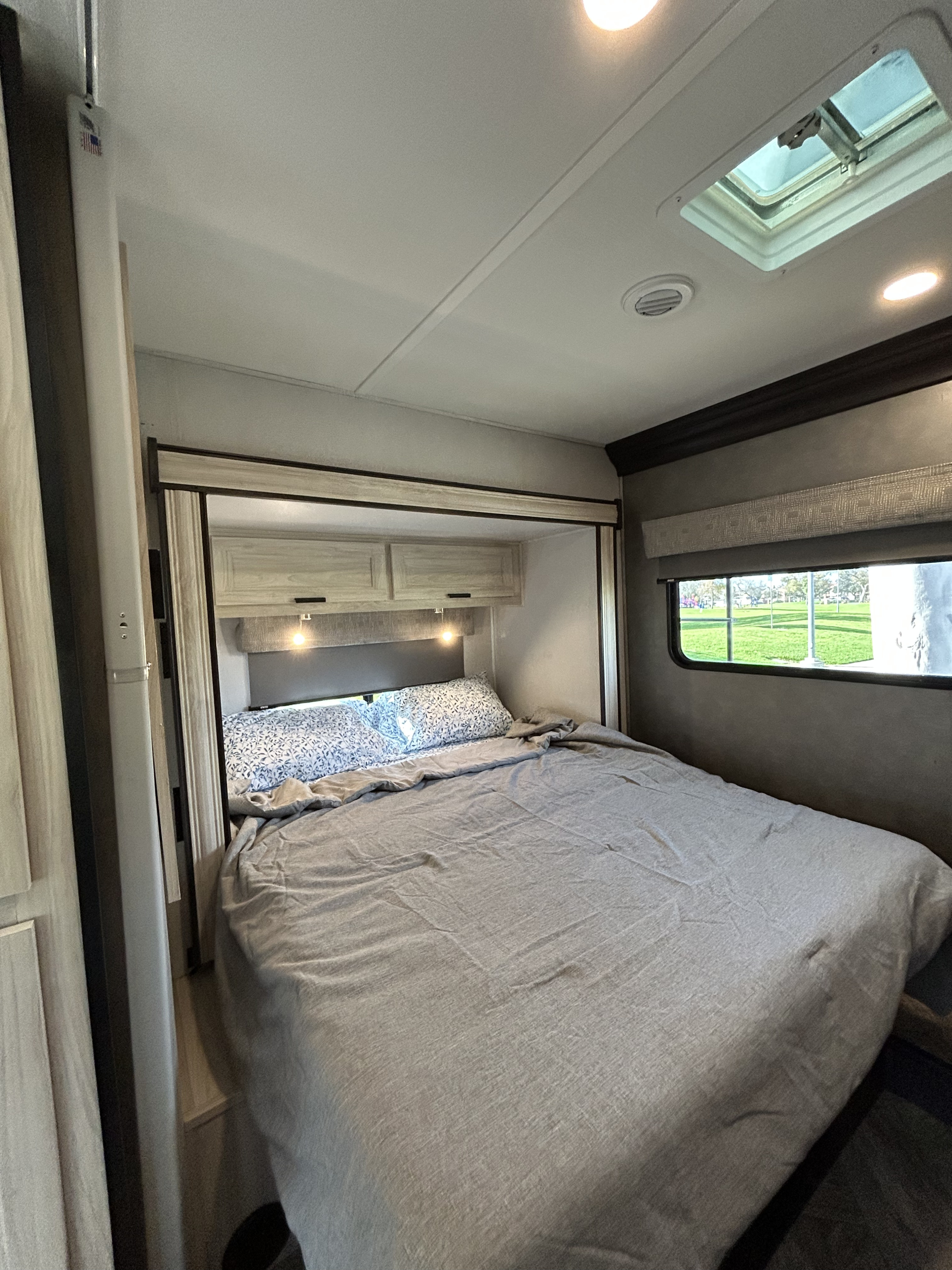 Inside an RV rental, this cozy bedroom features a comfortable bed, overhead cabinets, and a scenic window view.. Forest River Sunseeker Classic 2023