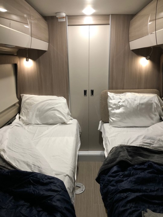 Cozy RV rental sleeping area featuring two twin beds with white linens, overhead storage, and a central closet.. Thor Sequence 2021