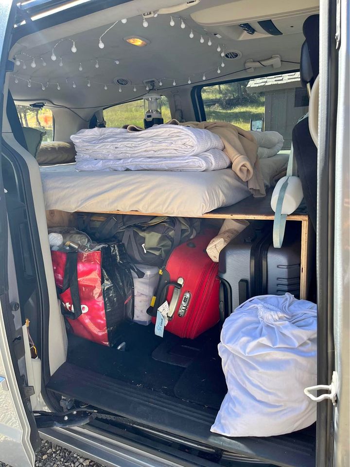 Inside an RV rental van: a custom bed platform with storage underneath, holding suitcases & bags. String lights adorn the ceiling.. Dodge Grand Caravan 2013