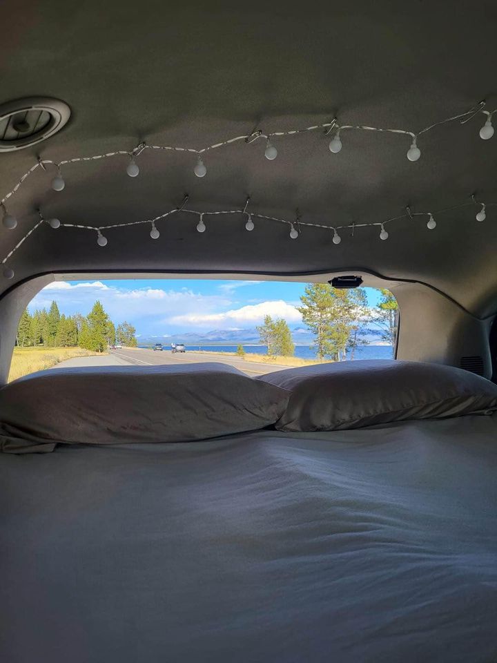 Inside an RV rental: a comfy bed with string lights, overlooking a scenic road, lake, and distant mountains. Perfect for van life travel.. Dodge Grand Caravan 2013