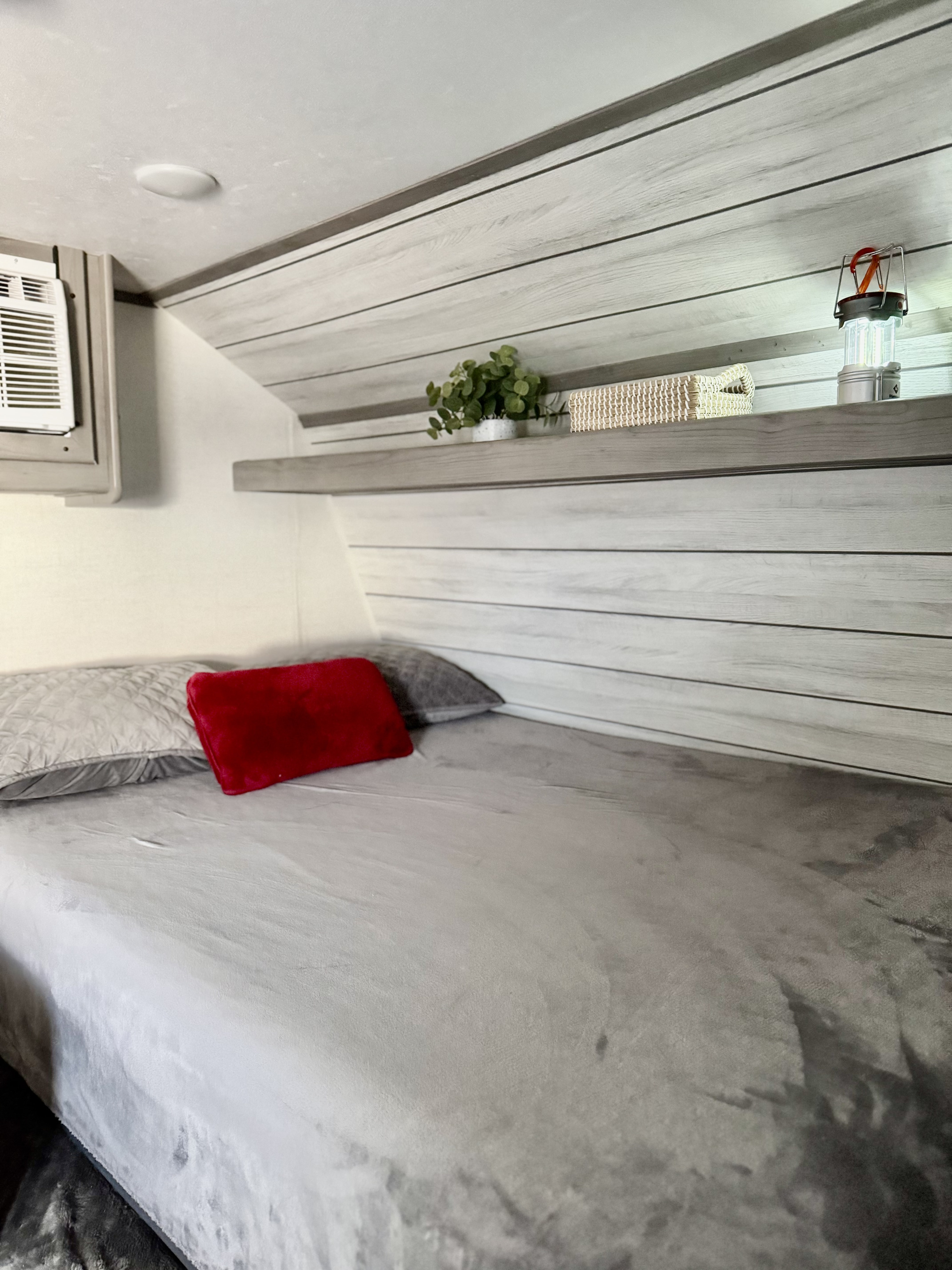 Cozy bed area in this RV rental, with grey bedding, a red pillow, and a decor shelf holding a plant and lantern.. Dutchmen Coleman Lantern LT 2023