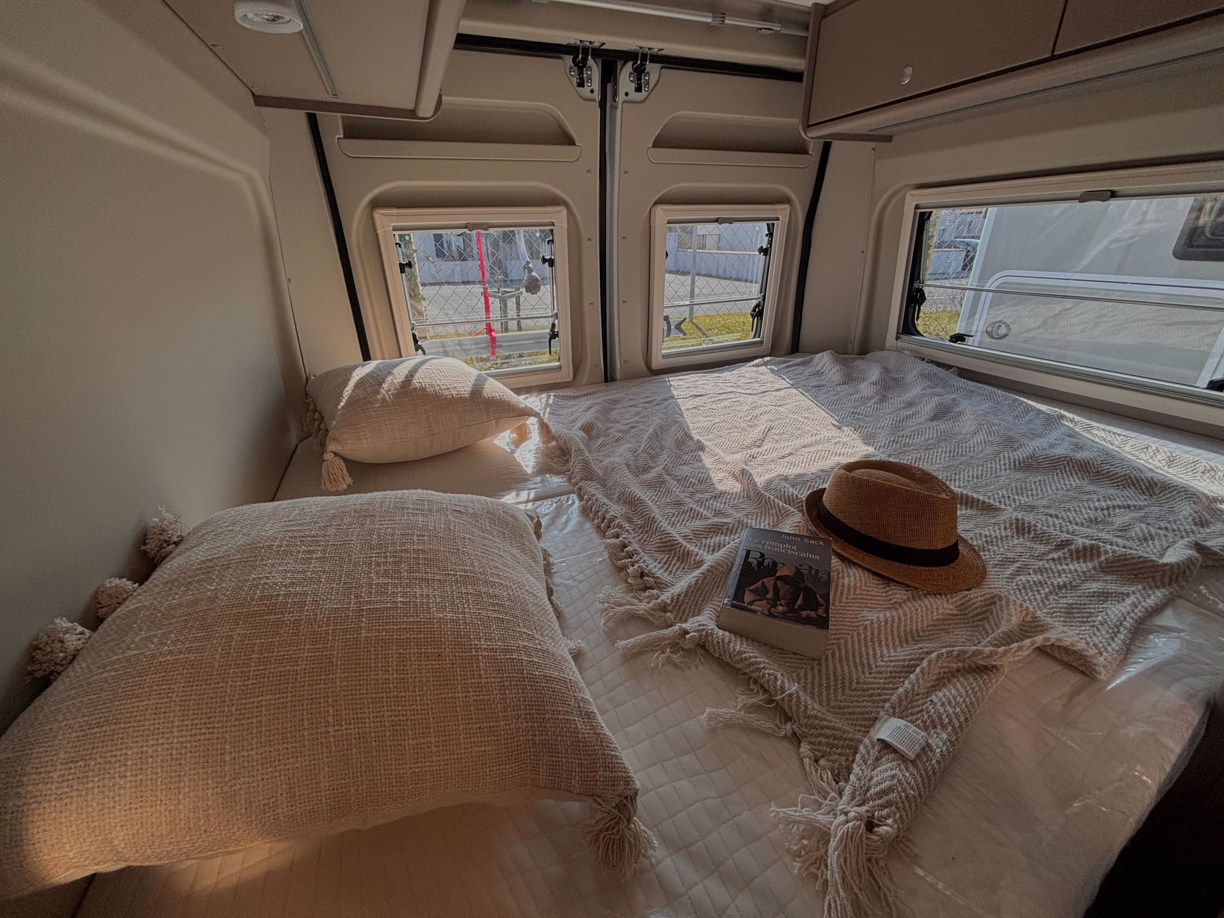 A serene RV rental bedroom, featuring a neatly made bed with soft pillows, a patterned blanket, a book, and a hat. Bright light streams through the rear windows.. CARADO CV-600 2024